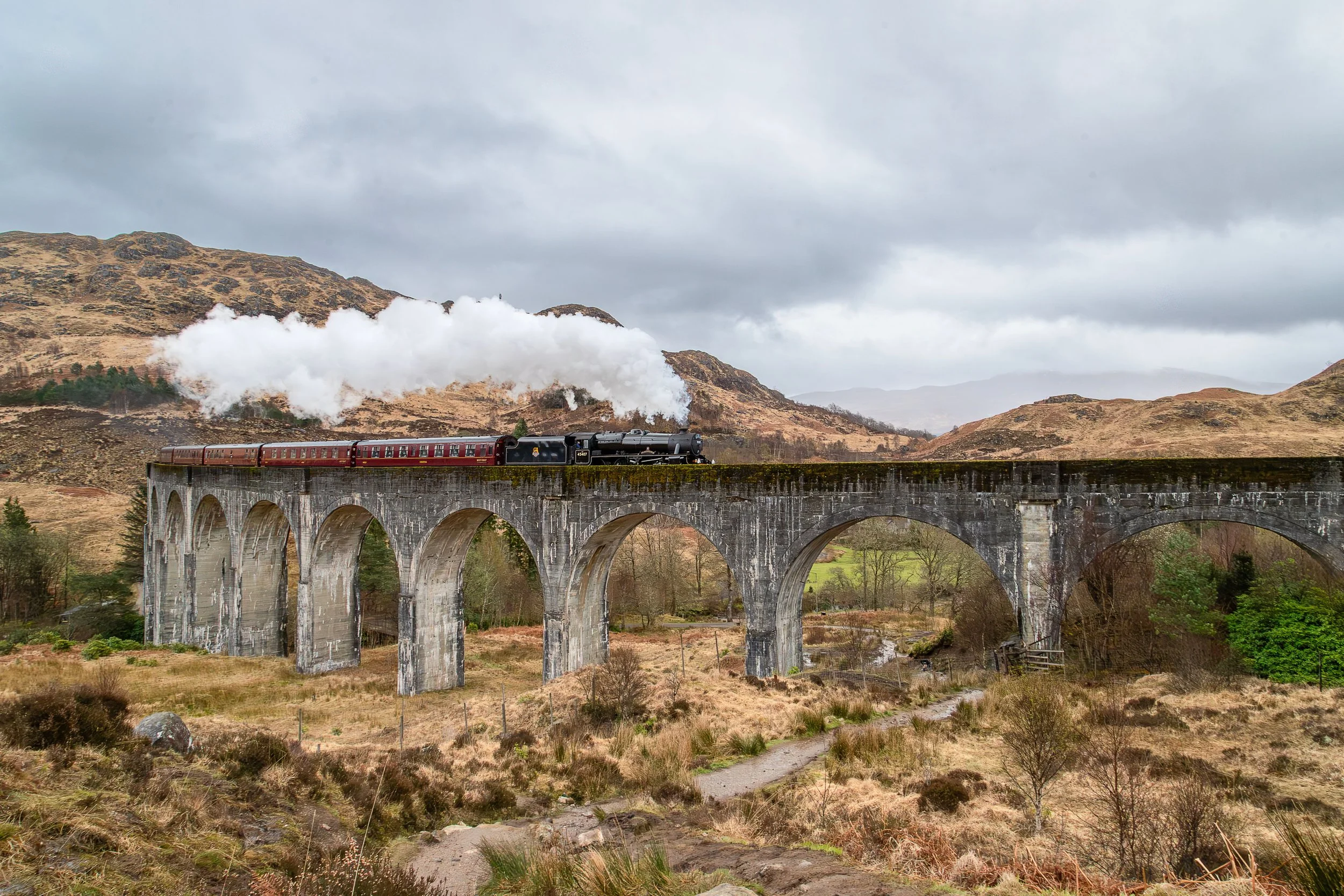 A vintage steam train traveling across a high stone arch bridge in a rural, mountainous landscape on a cloudy day.