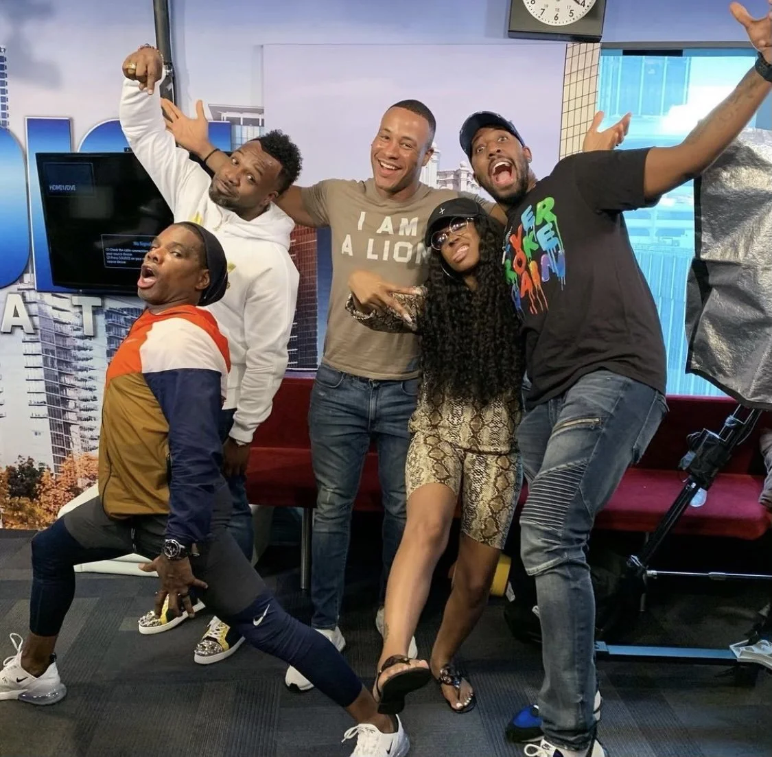 Five diverse people posing happily in a studio with a cityscape background, a TV screen behind them, and professional lighting equipment nearby.