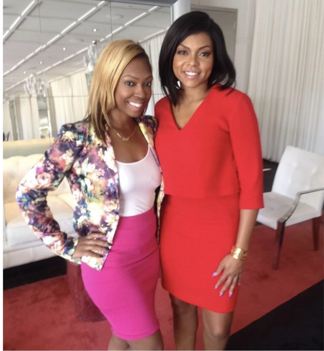 Two women standing close together, smiling, in a room with light-colored walls and furniture. One woman is wearing a floral blazer with a pink skirt, and the other is in a red dress.
