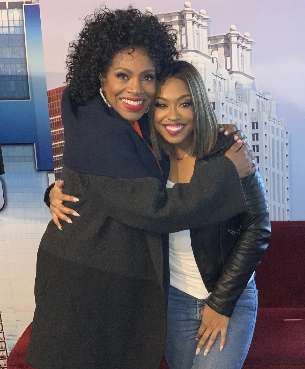 Two women hugging and smiling in front of a cityscape backdrop.