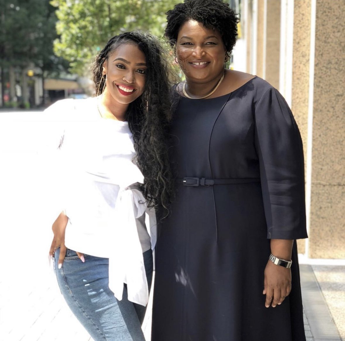 Two women standing close together outside, smiling at the camera. One woman is wearing a white top and jeans, and the other woman is wearing a dark dress.