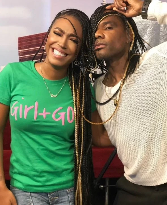 A smiling woman with long braided hair wearing a green shirt and a man with long braids wearing a white shirt posing together.