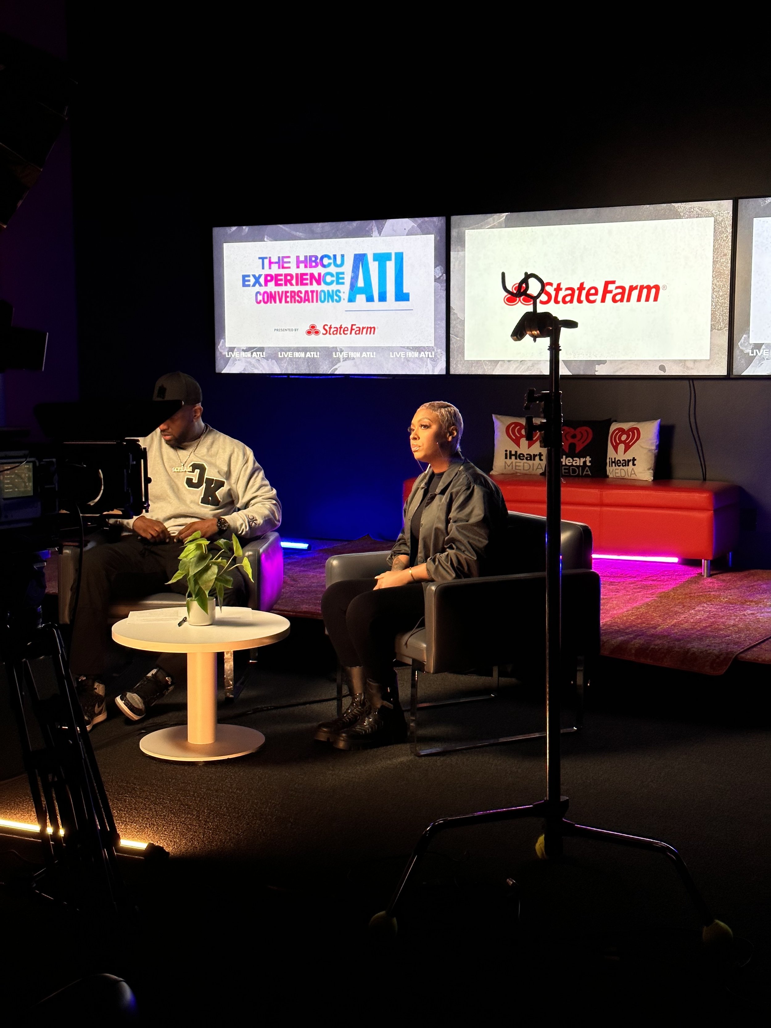 A television studio set with two large screens displaying the Atlanta Hawks Center of the Atlanta Hawks basketball team, with the logos of State Farm Insurance and iHeartMedia. There are two seated hosts, one male and one female, with studio equipmen