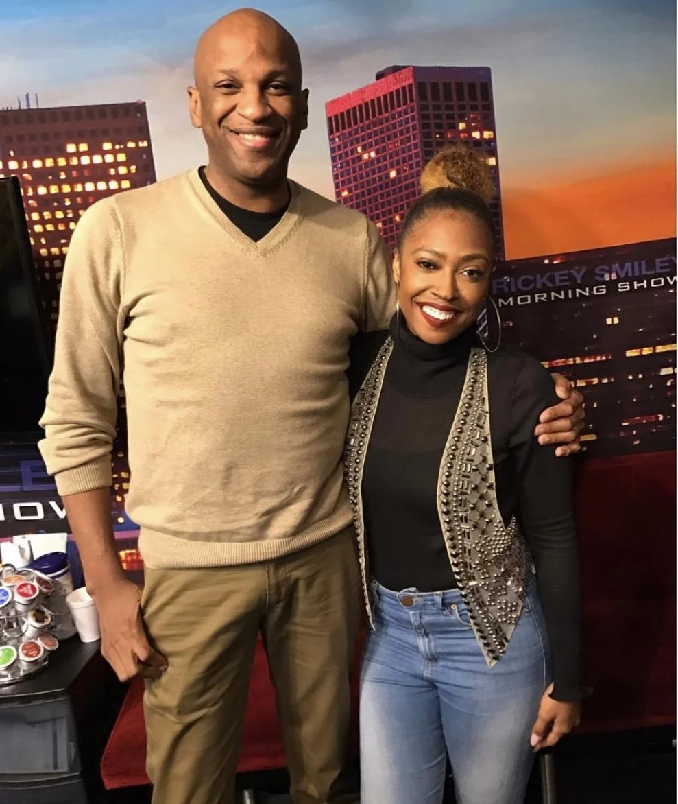 A man and a woman smiling, standing close together with an urban cityscape backdrop at sunset. The man is wearing a beige sweater and khakis, and the woman is wearing a black top with an embellished vest and light blue jeans.