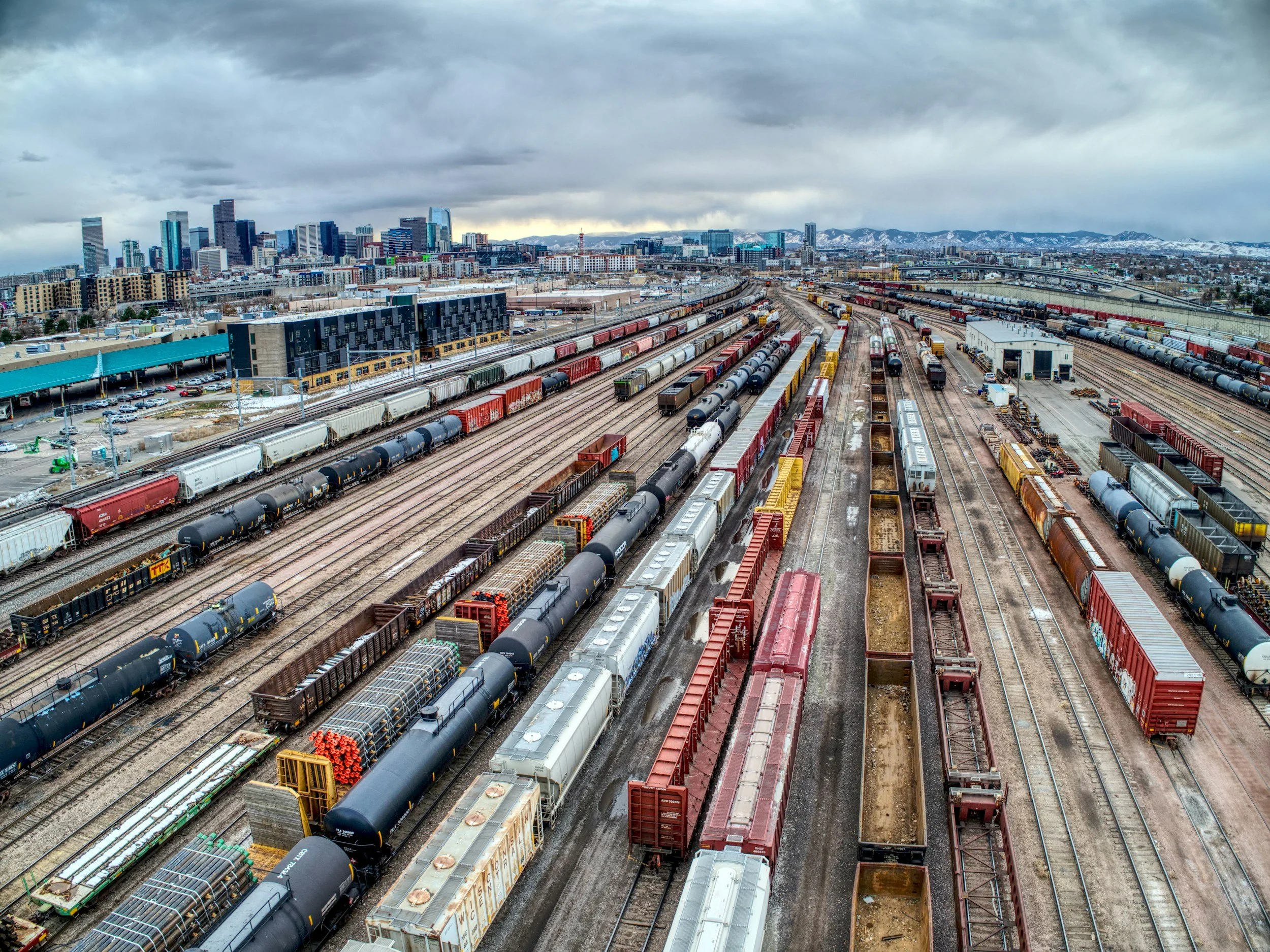 Aerial view of a freight train yard with multiple train tracks filled with various freight cars and a city skyline with tall buildings in the background under a cloudy sky.