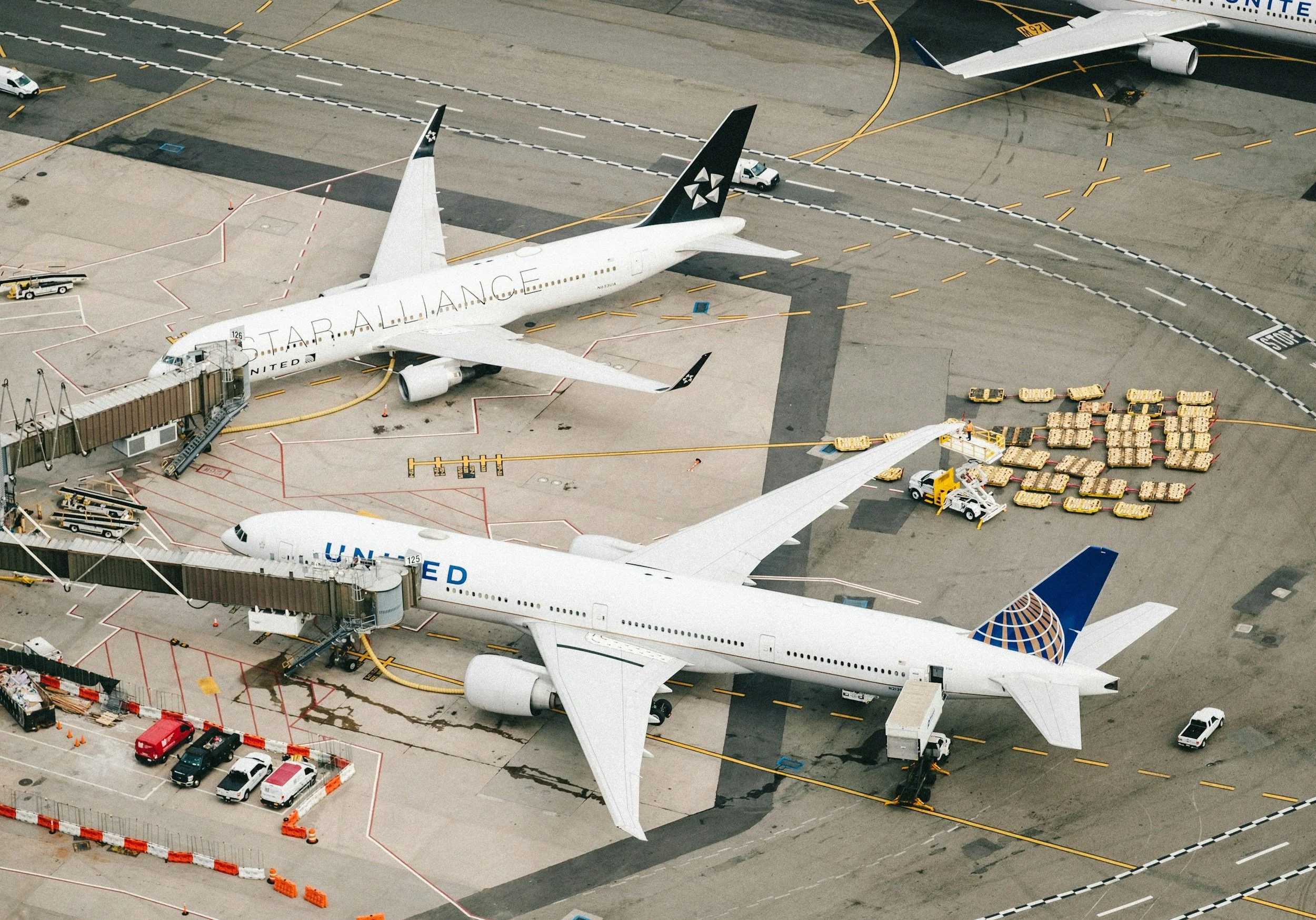 Aerial view of airport tarmac with multiple airplanes, ground vehicles, and cargo pallets. One airplane has the words 'Star Alliance' on its side.