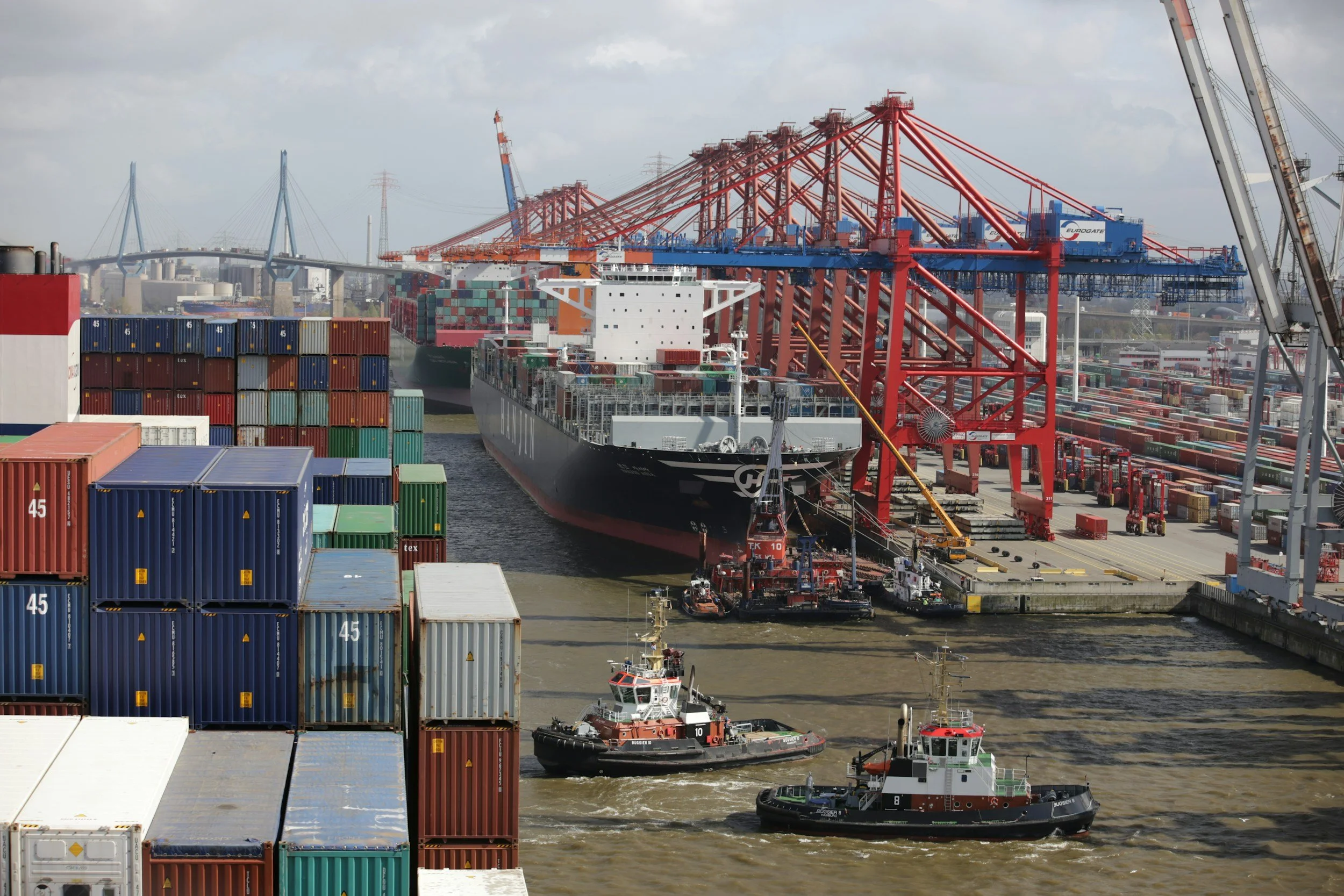 A busy shipping port with container ships, colorful stacked containers, and large cranes moving containers around. Tugs guide ships in the harbor, with a cityscape and bridge in the background.