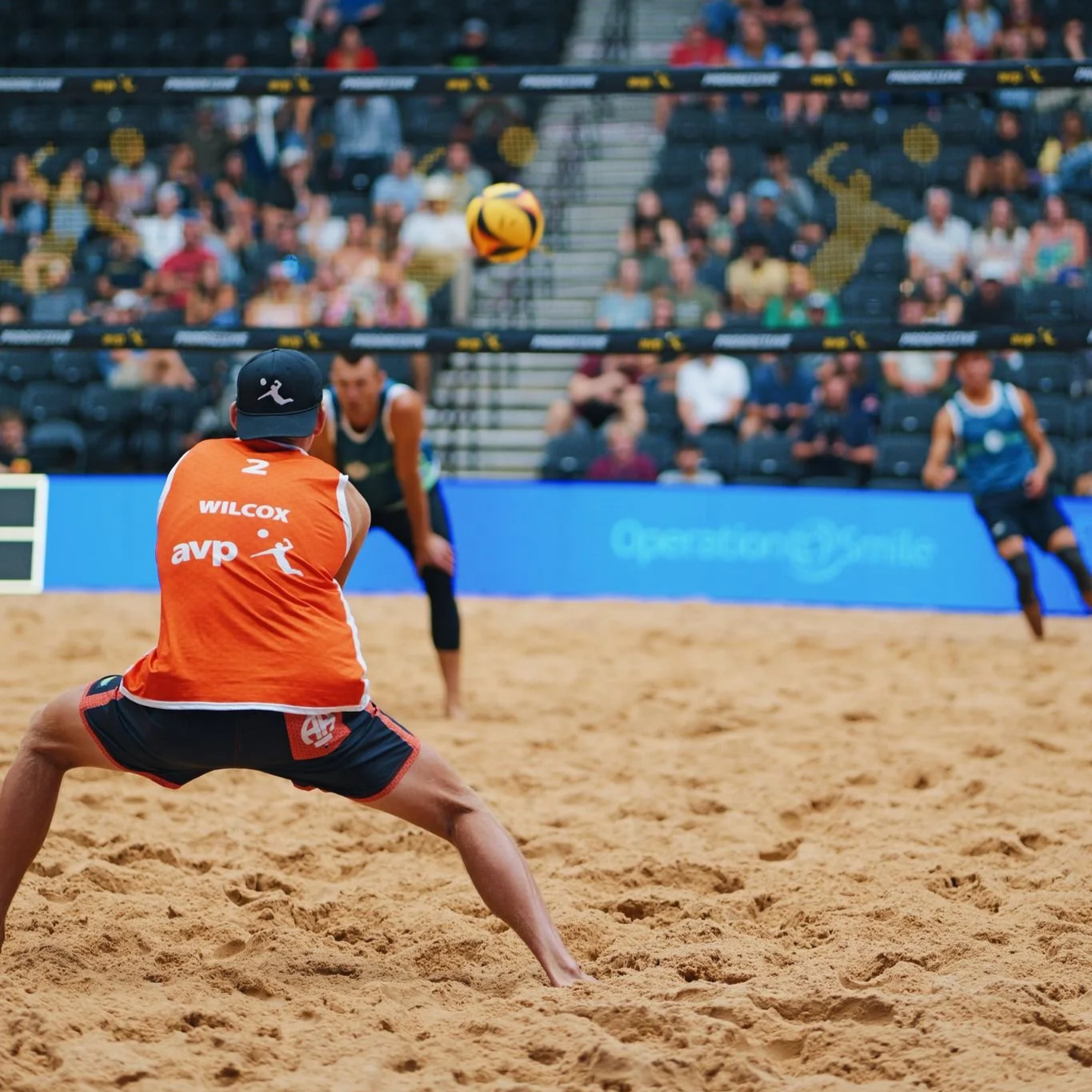 A beach volleyball game in progress with players and a ball, crowd in the background.