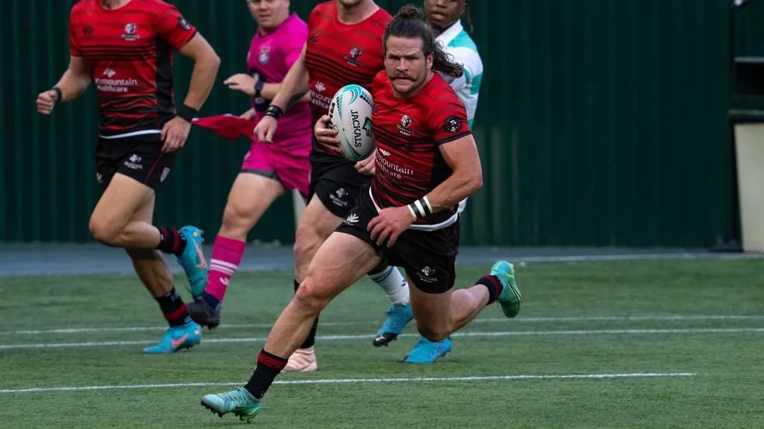 A rugby player with long hair and a mustache running with the ball, wearing a red and black jersey, black shorts, and teal shoes, on a rugby field with teammates and opponents in the background.