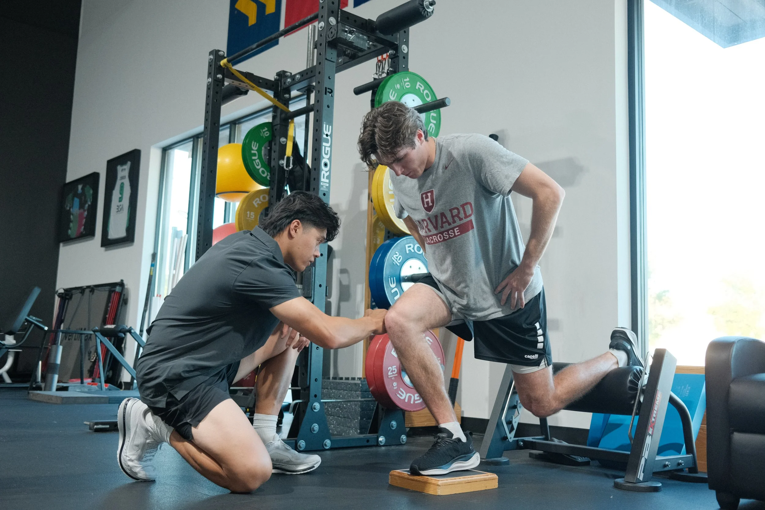 A personal trainer assists a young man during a weightlifting exercise on a leg curl machine in a gym, with weight plates and gym equipment visible in the background.