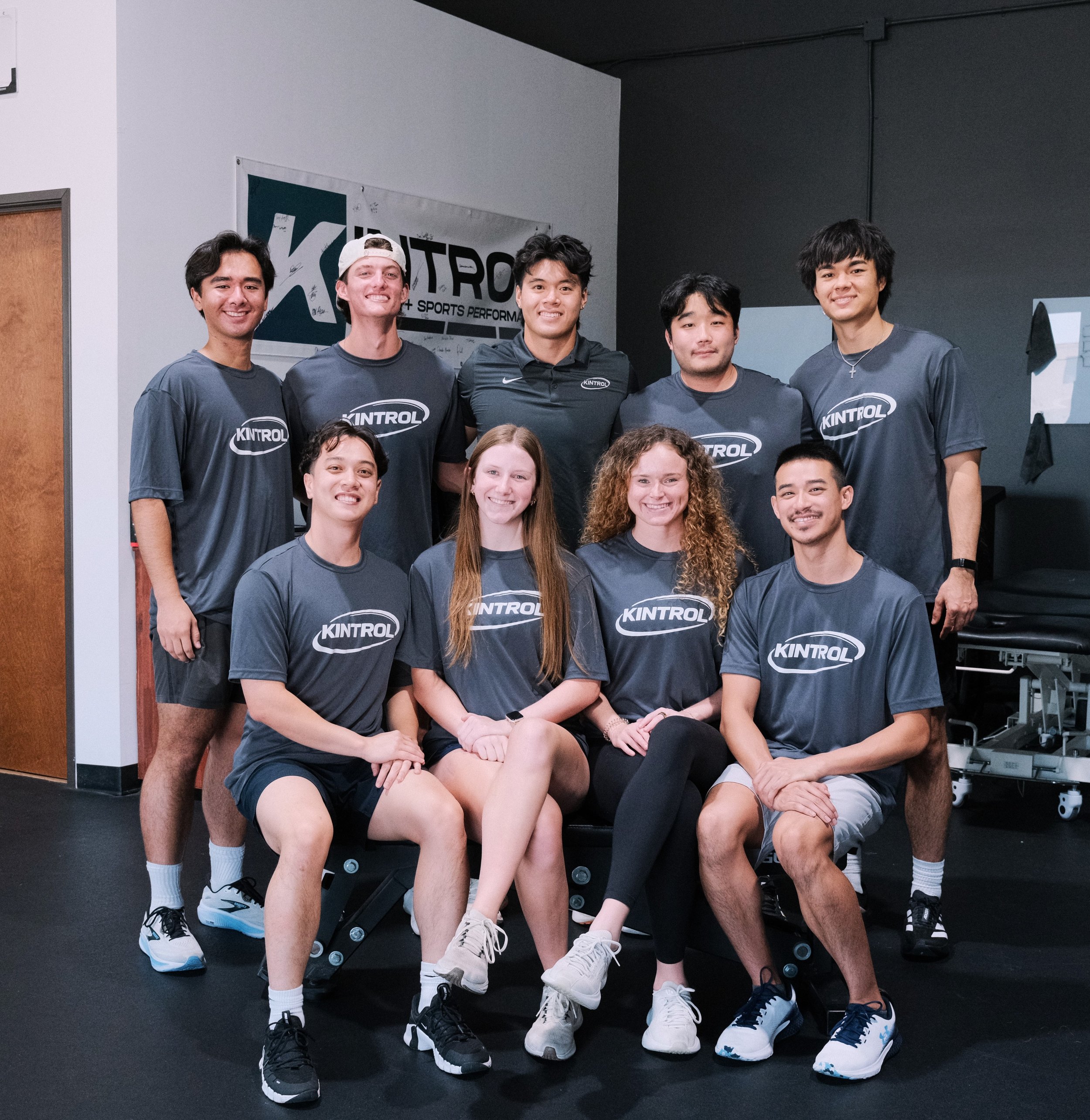 Group of young athletes wearing gray Kintrol sports shirts, sitting and standing in a gym, smiling for a team photo.
