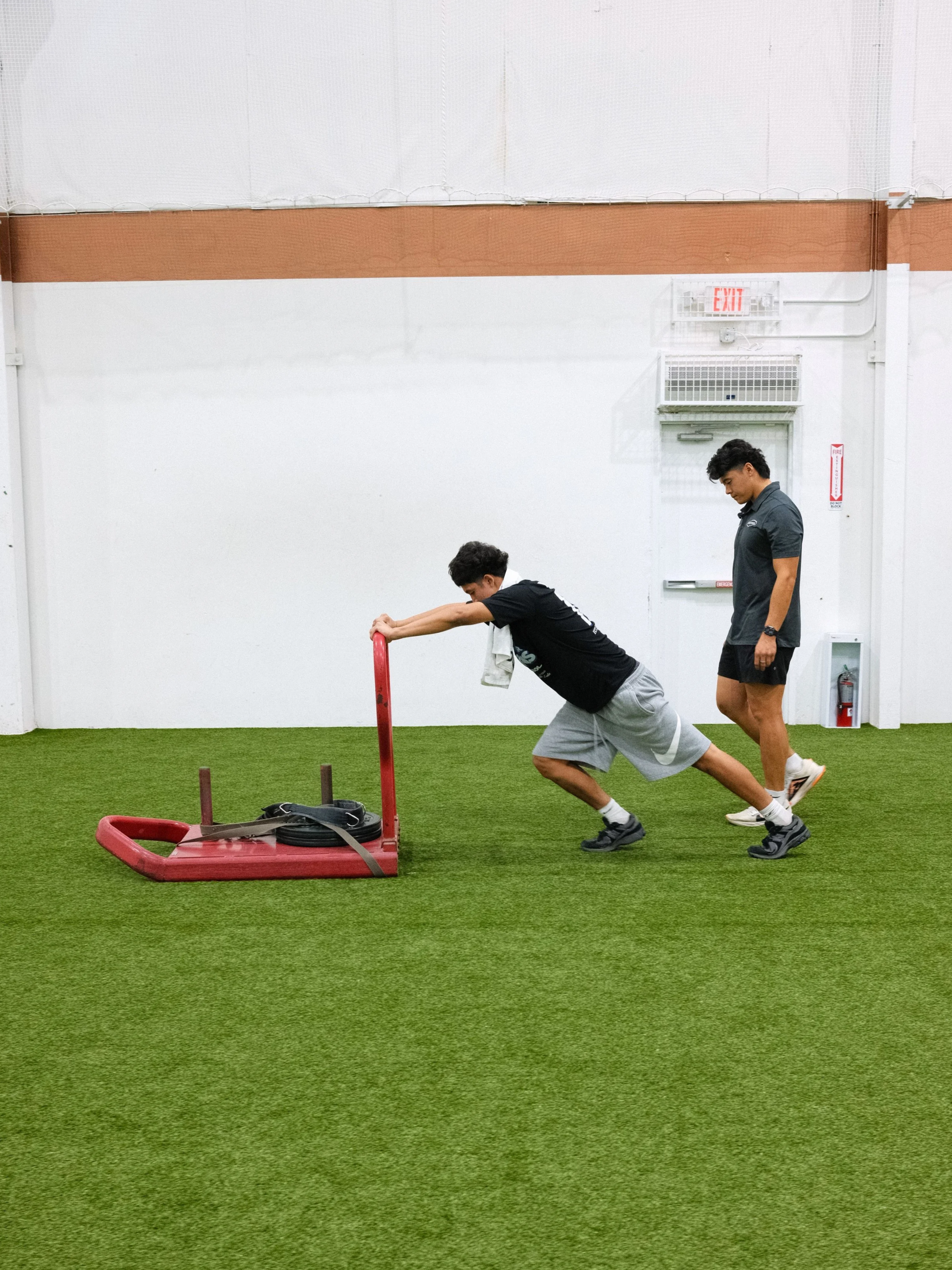Two athletes pushing a weighted sled across indoor turf during a gym workout.