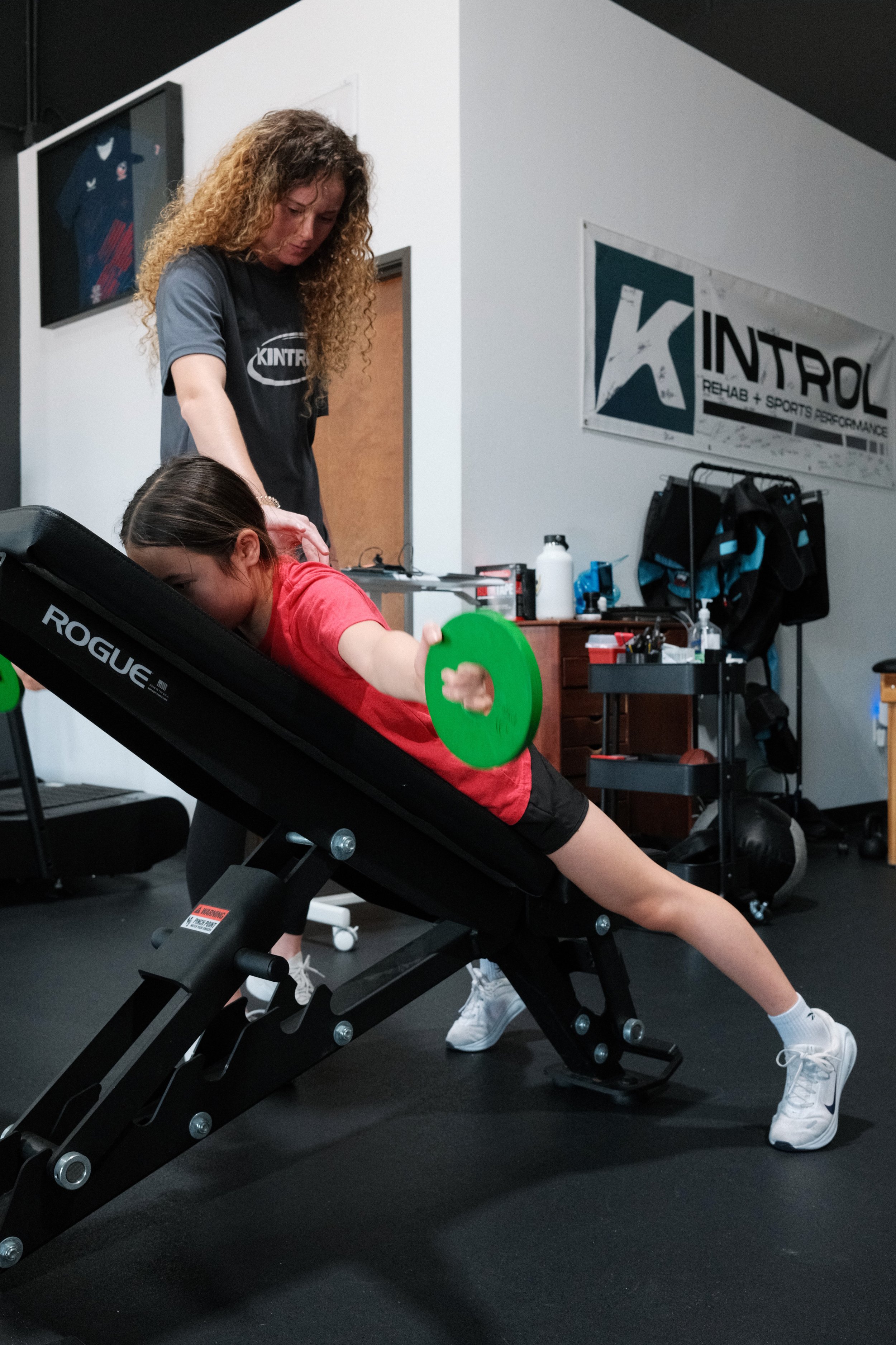 A young woman with brown hair in a ponytail is performing a stretching exercise on a black adjustable workout bench in a gym. She is reaching forward, holding a green weight plate, with her left leg extended straight back and her right knee bent. An instructor, a woman with curly brown hair, is standing behind her, guiding her exercise. The background includes a white wall with a gym banner that reads 'KINTROL REHAB + SPORTS PERFORMANCE', and various fitness equipment such as a water bottle, a backpack, and weights.