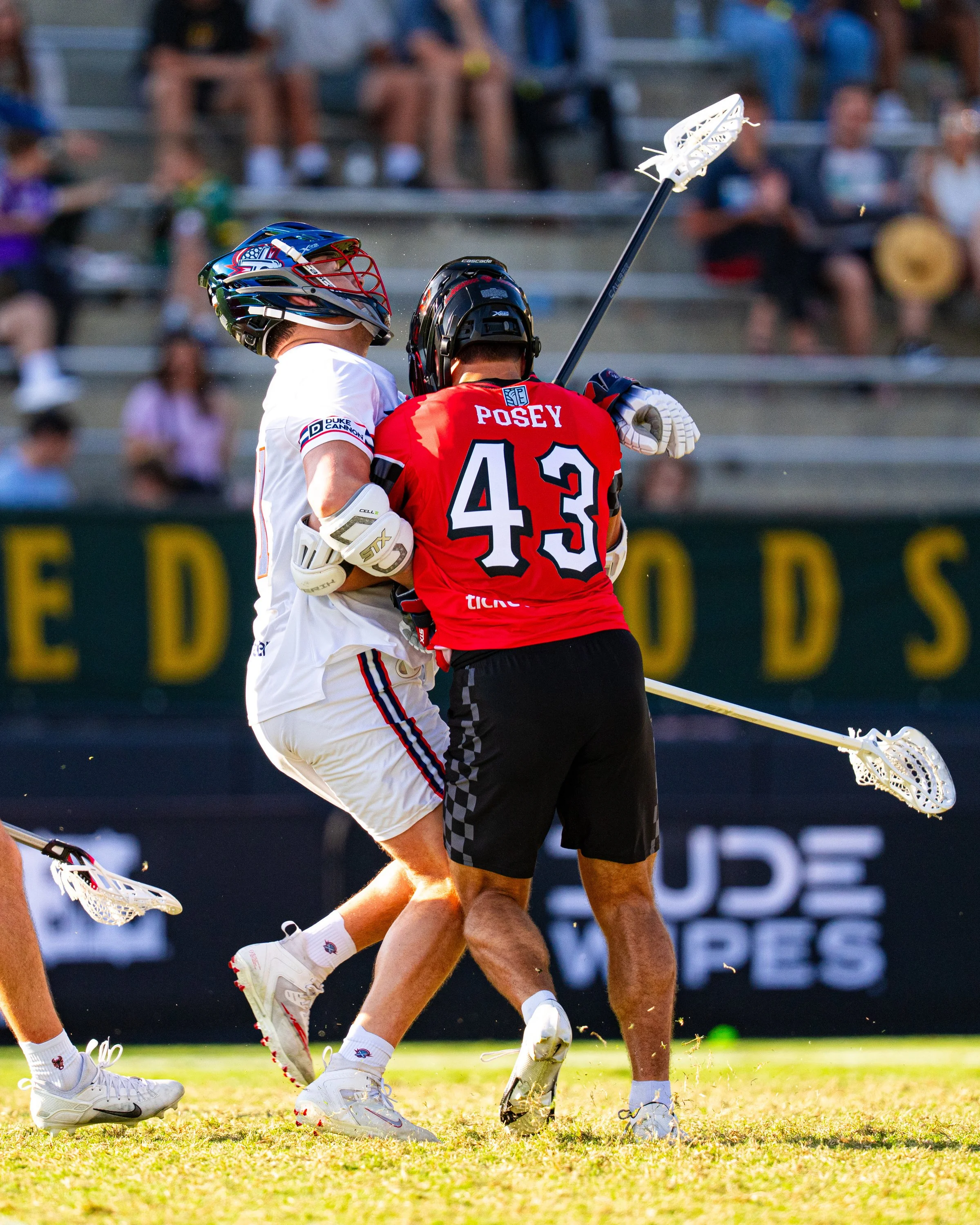 Two lacrosse players competing for the ball during a game, with a background of spectators sitting in stands.