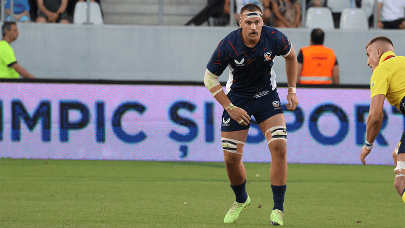 A rugby player in a navy uniform with white and yellow accents and bandages on his knees and arm, running on the field during a match.
