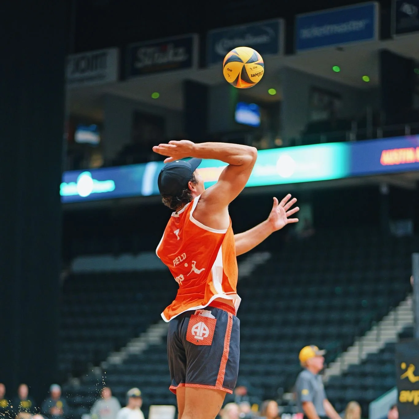 A person in an orange sports jersey and gray shorts is playing indoor volleyball, preparing to serve the ball, with their right arm raised and the ball in the air.