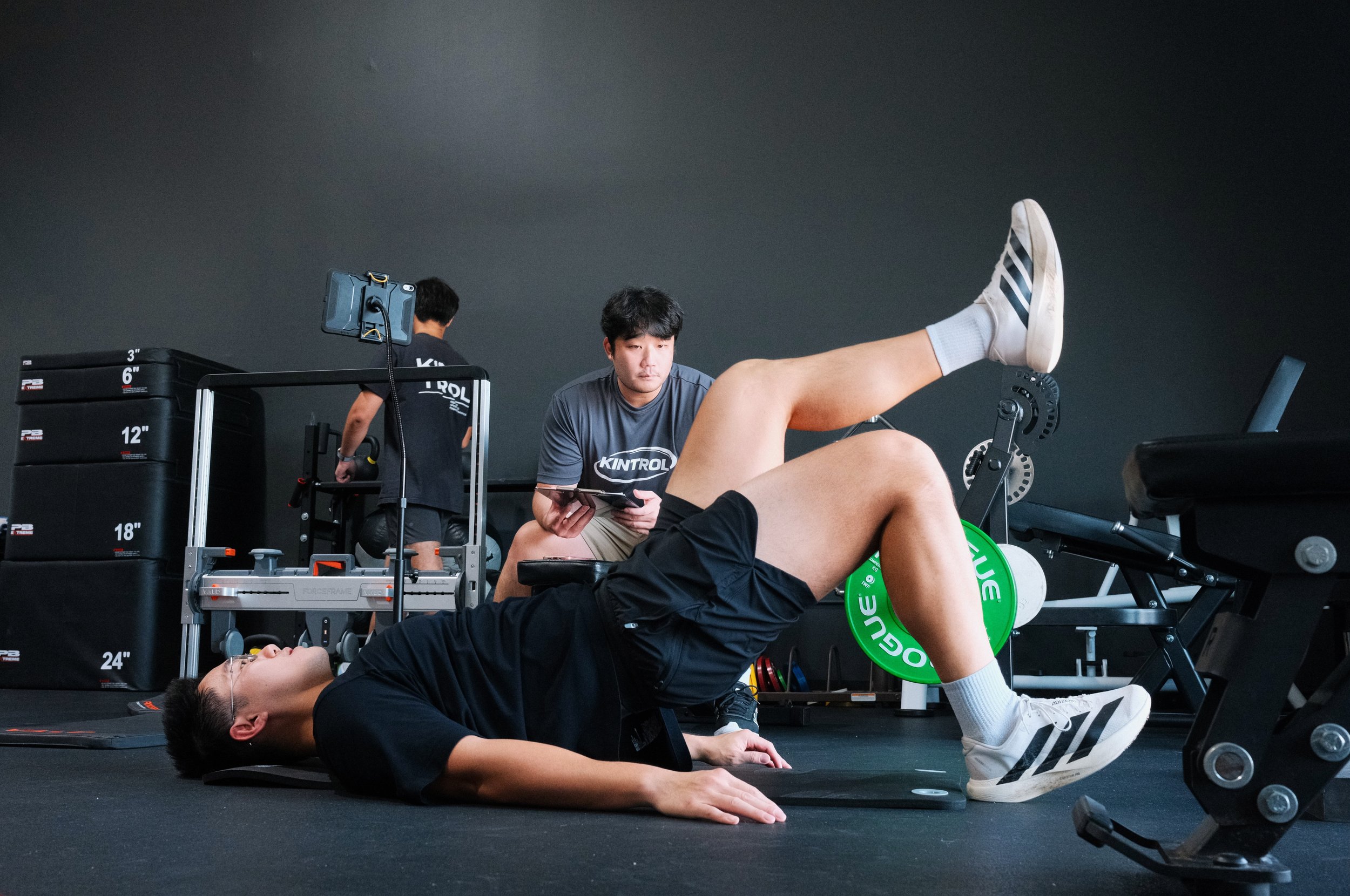 A fitness trainer assisting a client during a workout session in a gym, with gym equipment visible and a black wall in the background.