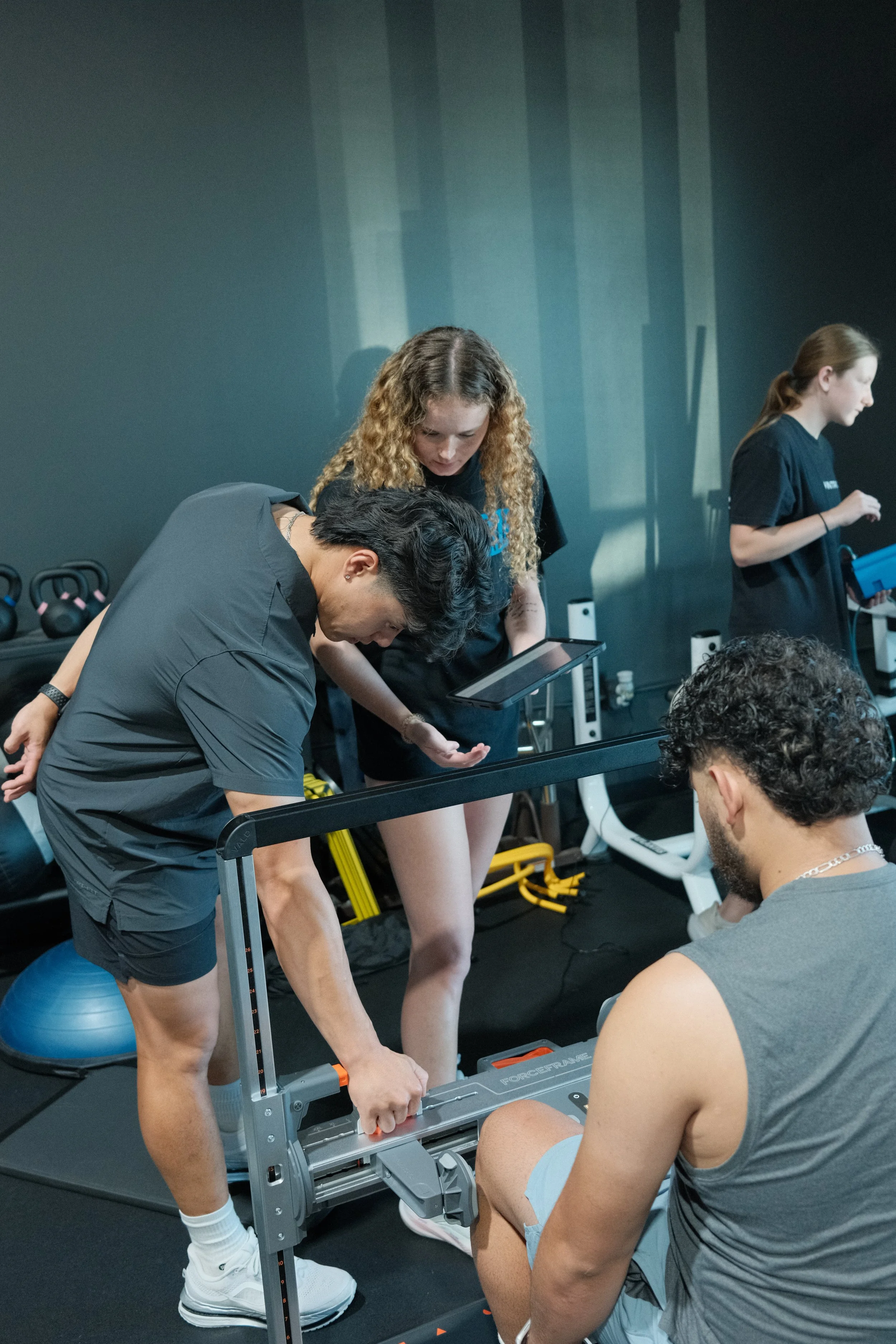 Group of people at gym engaging in a fitness activity with a focus on a woman adjusting a piece of gym equipment. Others are seen working out or preparing in the background.