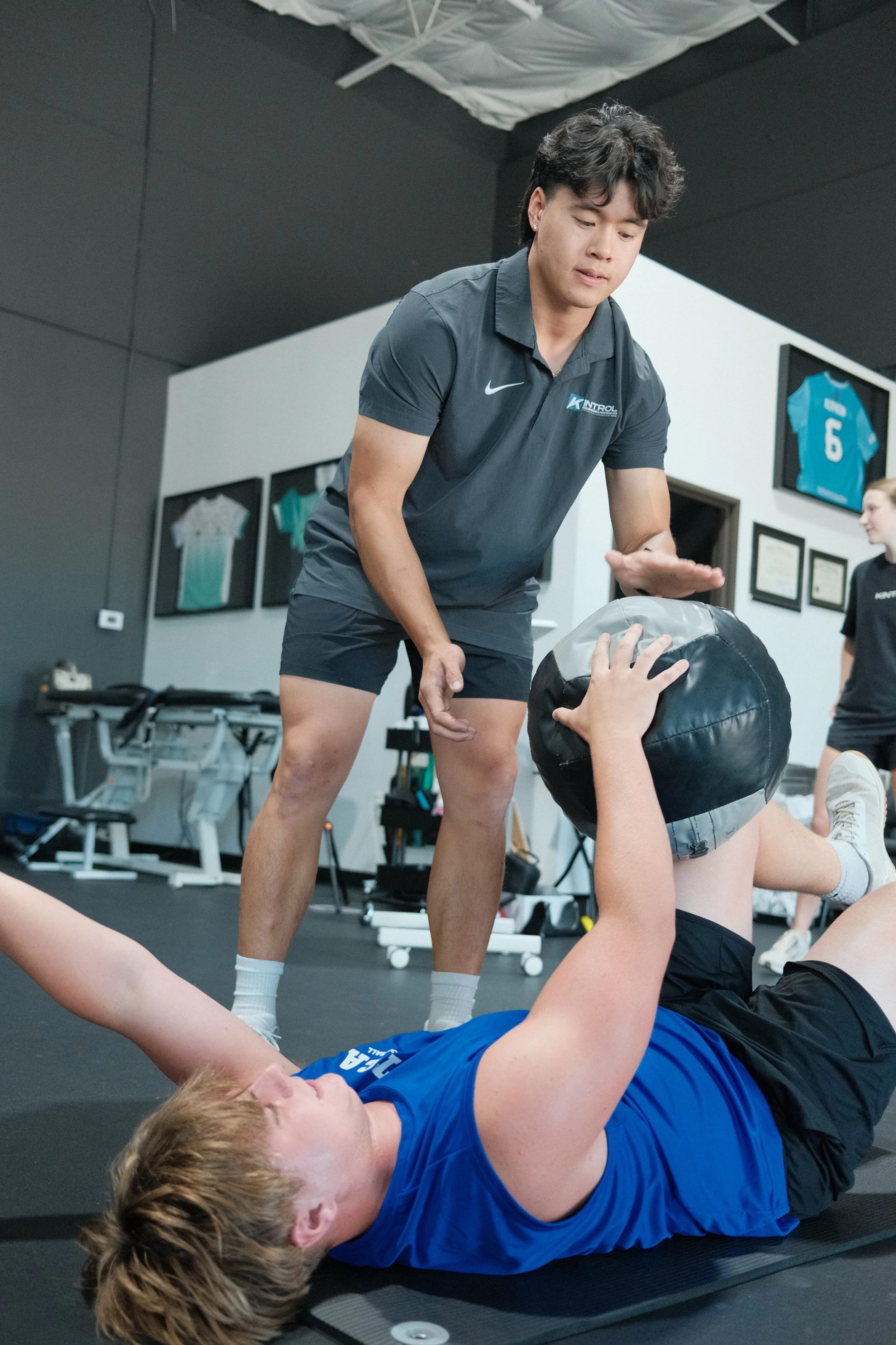 A young man in a navy polo shirt assists a young woman lying on a workout mat, holding a medicine ball with her legs raised. The woman is wearing a black sports bra and shorts. There are framed sports jerseys on the wall in the background, indicating a sports training or therapy setting.