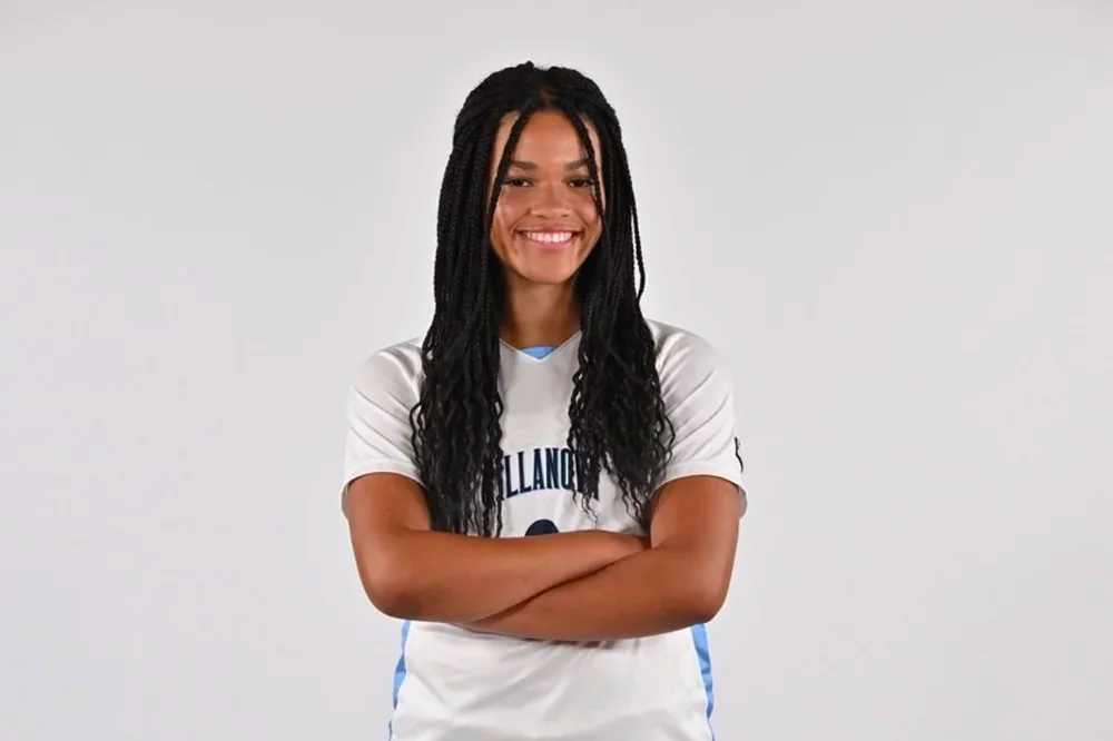 A young woman with long braids smiling, standing with arms crossed in a white sports jersey with blue accents.