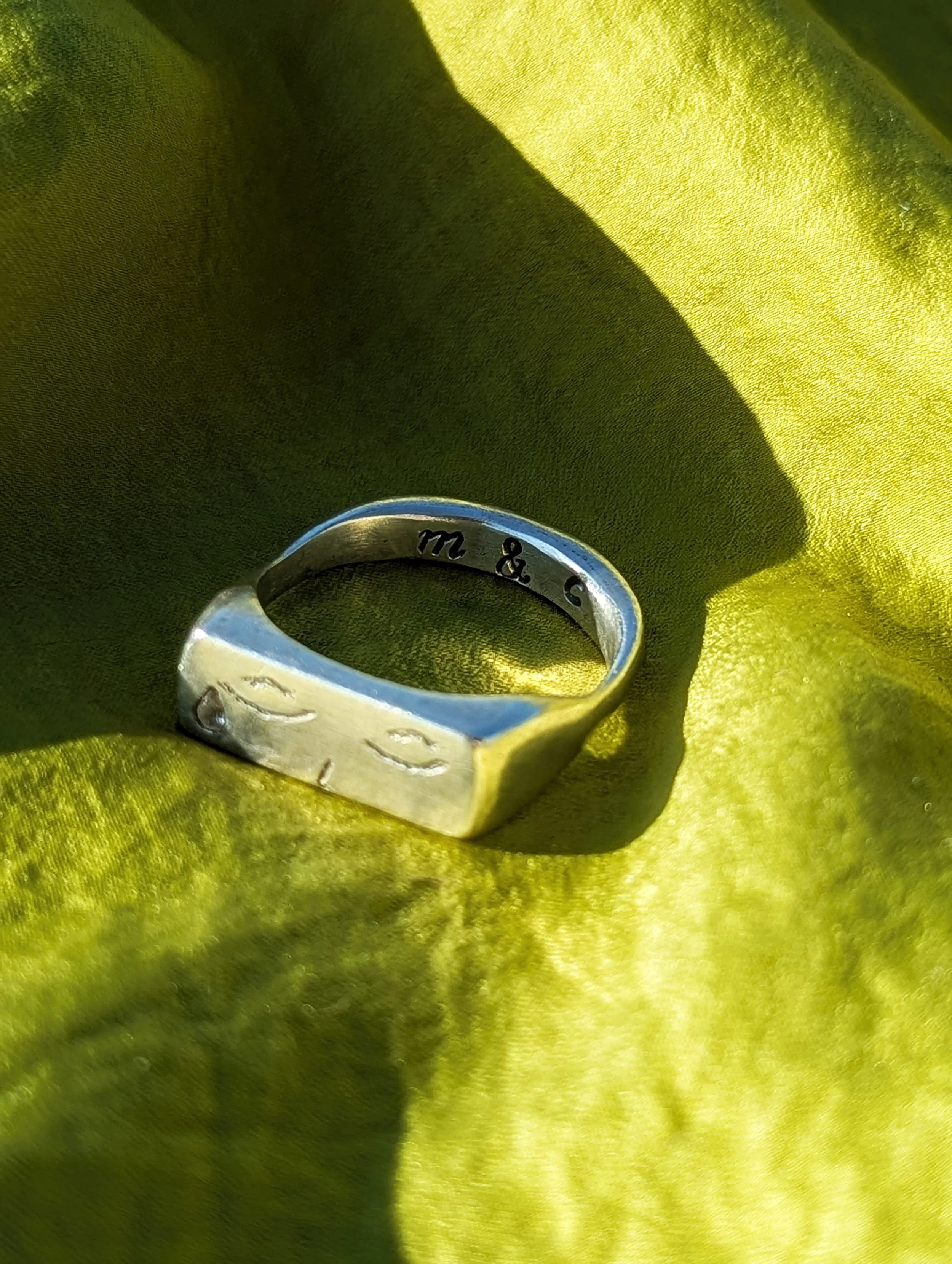 A close-up of a silver ring with engraved initials 'M' and 'C' placed on a yellow-green fabric surface.
