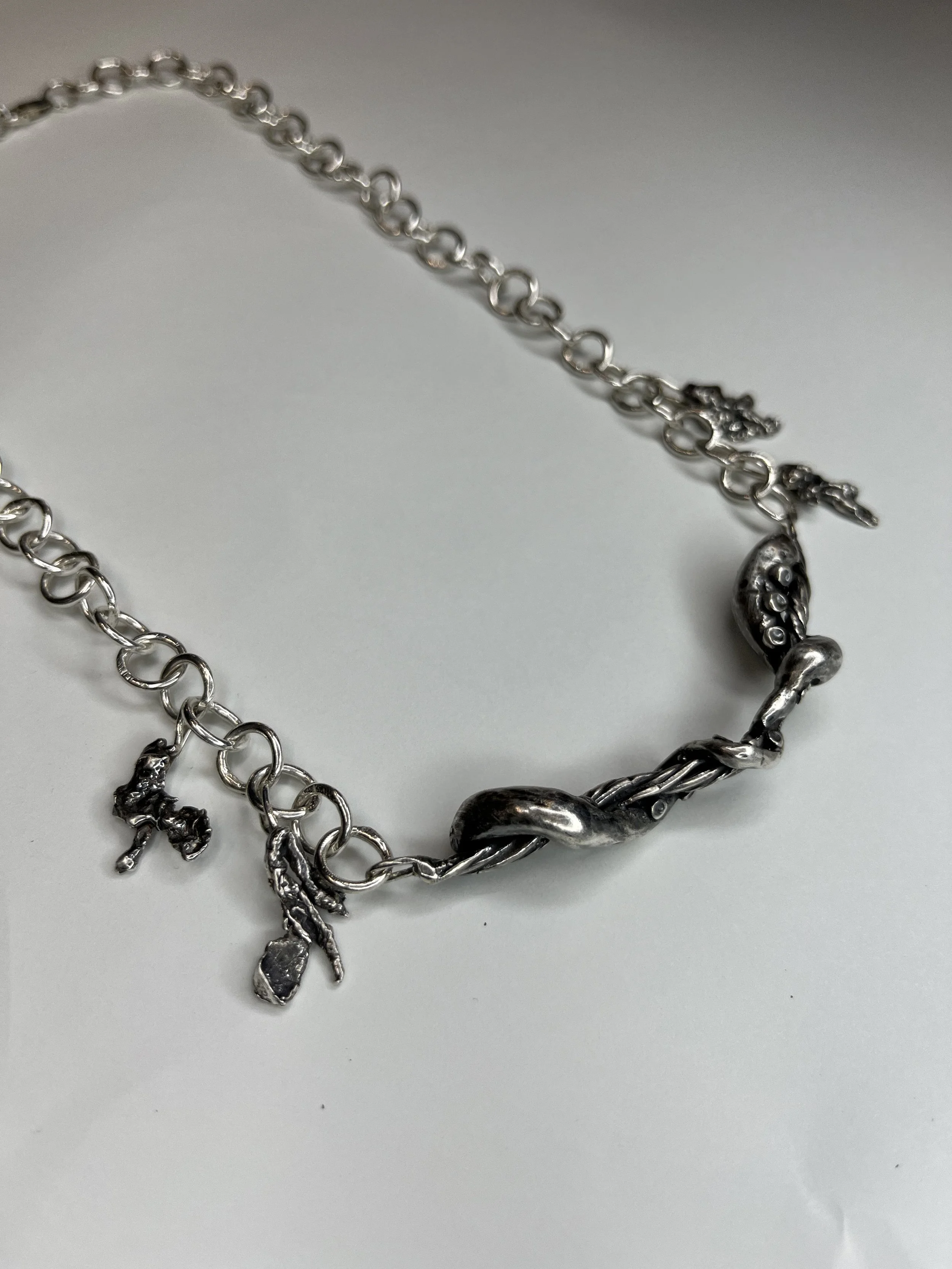 Silver necklace with various charms, including a shoe, hand, and a small animal or figure, on a white background.