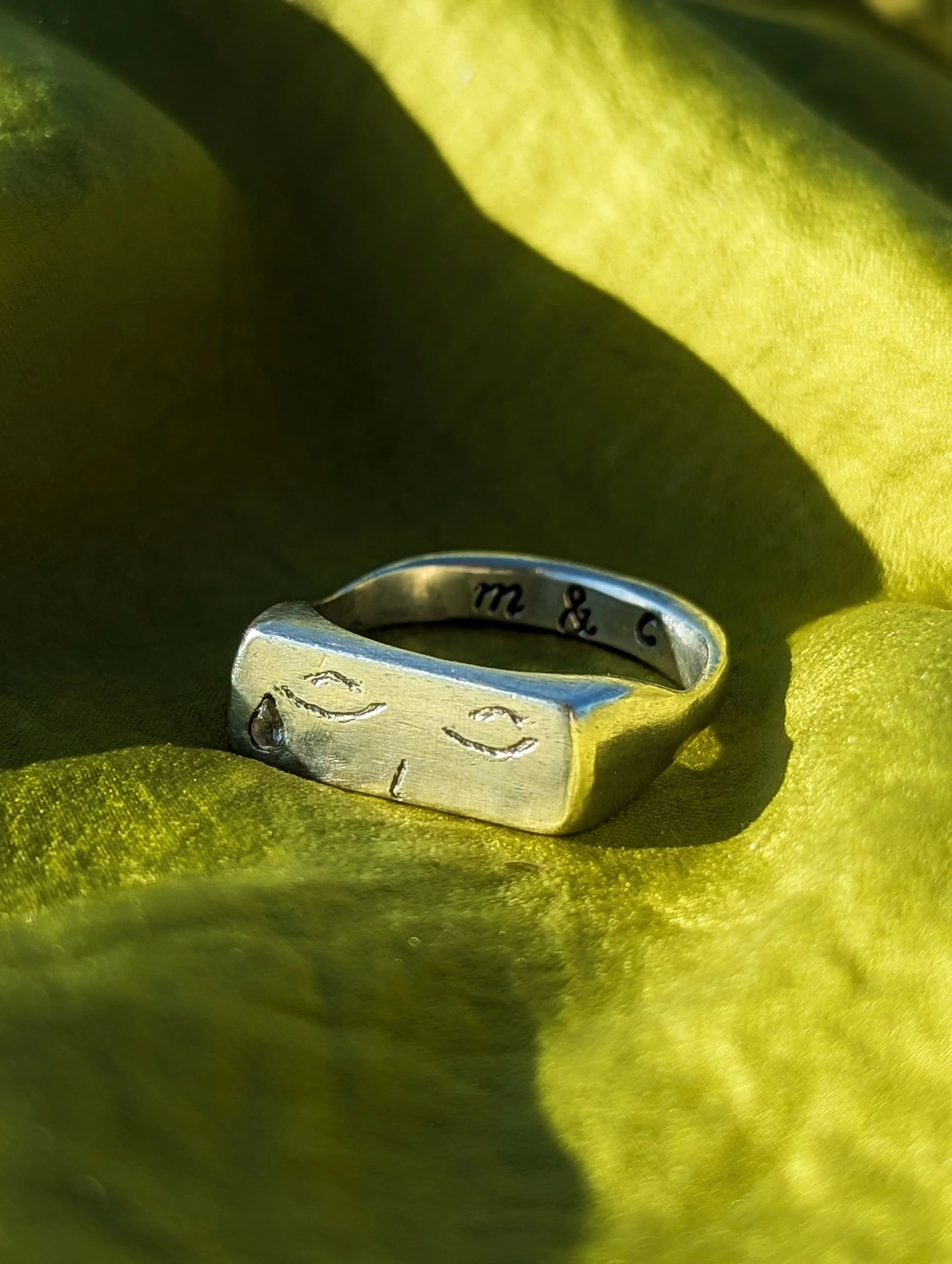 A close-up of a silver ring with engraved initials 'm & c', an engraved face with a tear drop, placed on bright green fabric.