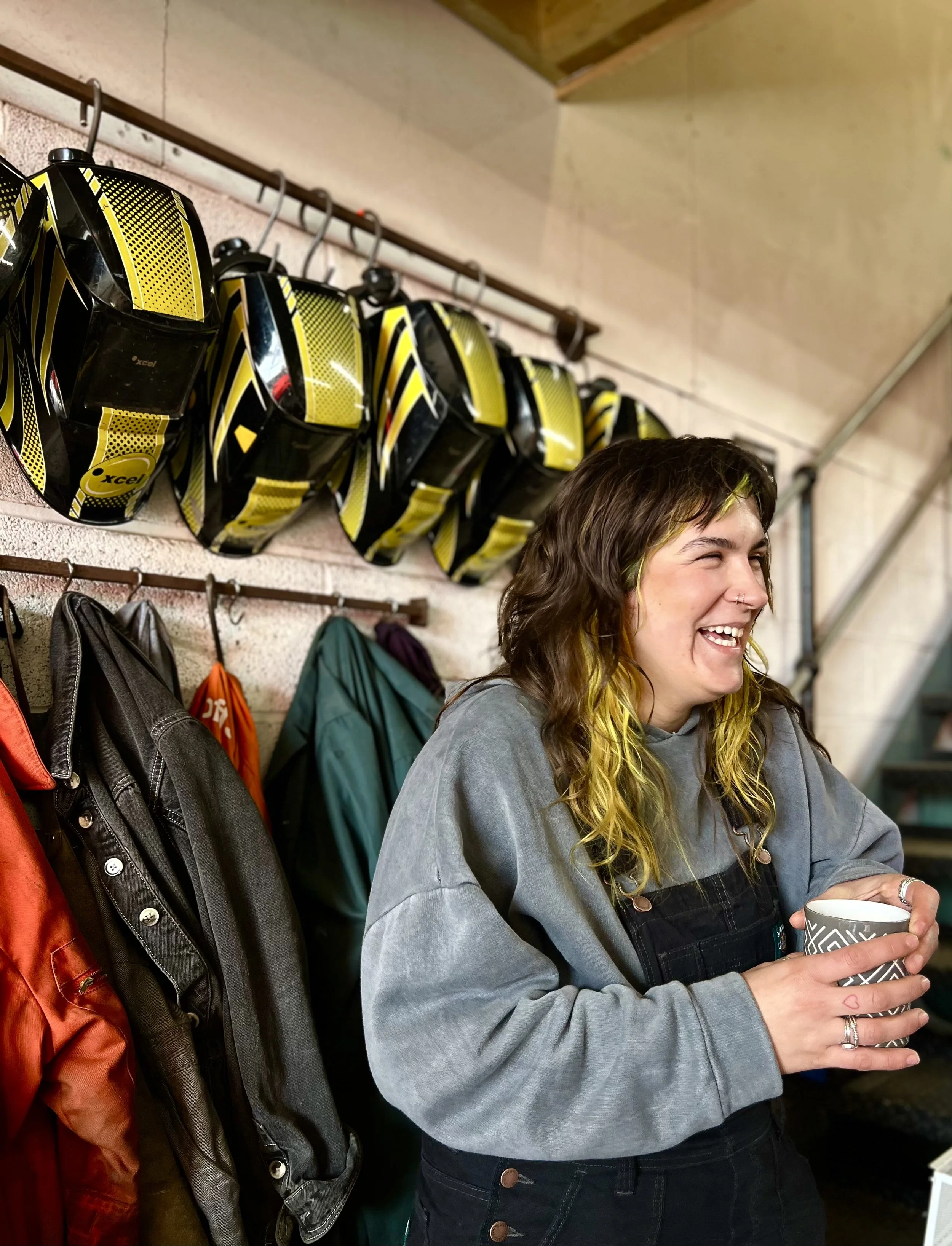 Young woman with brown and yellow hair smiling and holding a patterned coffee mug, standing in front of a row of yellow and black bicycle helmets hanging on a wall, with backpacks on a rack below.