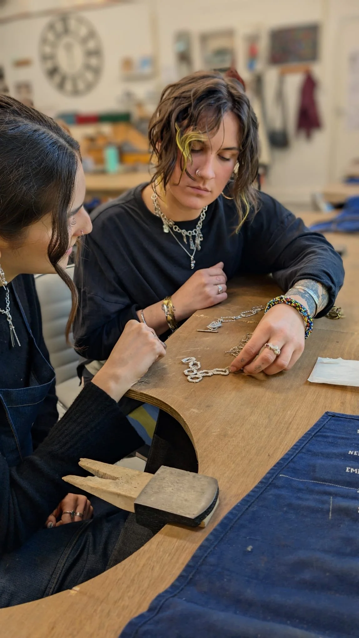 Two women are sitting at a wooden table, examining jewelry. One woman has short, wavy hair with yellow streaks, multiple necklaces, and rings. The other woman has long dark hair and also wears rings and an earring. Jewelry and tools are on the table, including a hammer and jewelry parts. The background is a workshop with shelves and wall decorations.