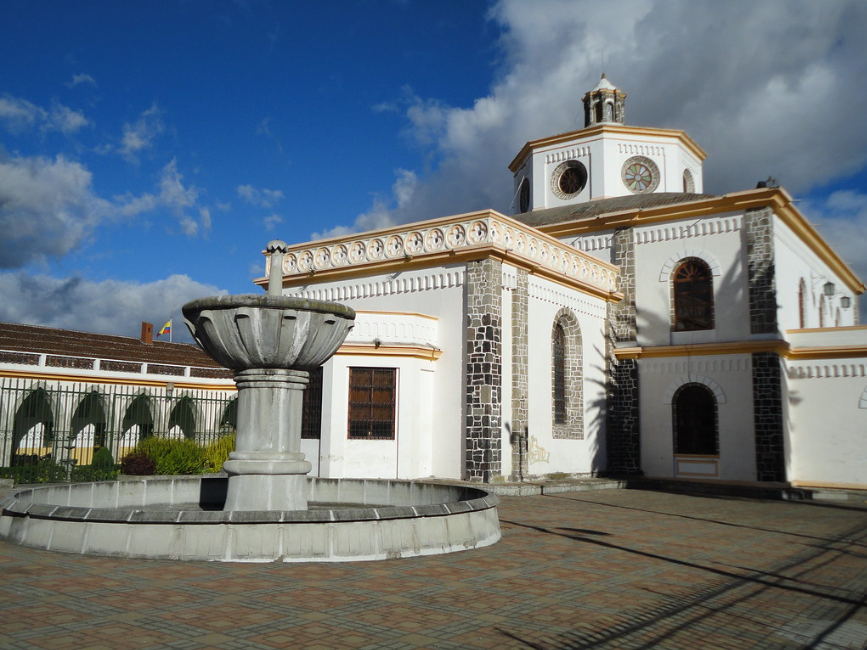Iglesia antigua de piedra y yeso blanca con reloj en la torre, fuente de piedra en primer plano, cielo con nubes, en un día soleado.