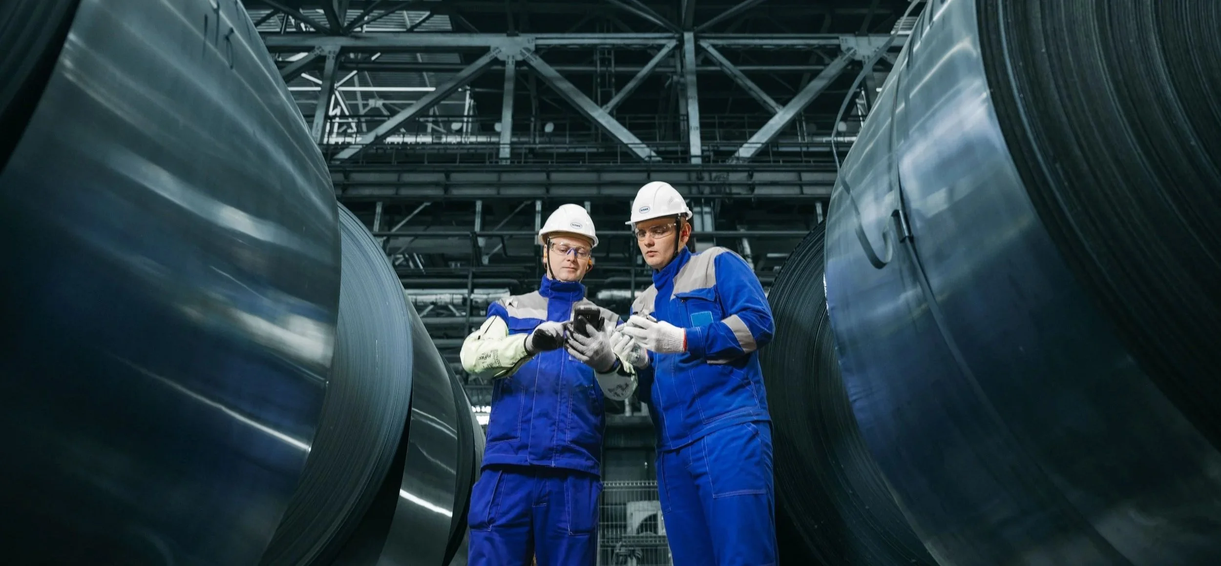 Dos trabajadores en uniforme de seguridad con cascos revisando información en un teléfono en una fábrica o planta industrial rodeada de grandes bobinas de metal.
