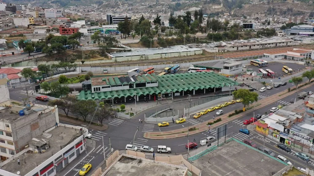 Vista aérea de una estación de autobuses en un área urbana, rodeada de calles con autos y taxis, y edificios en el fondo.
