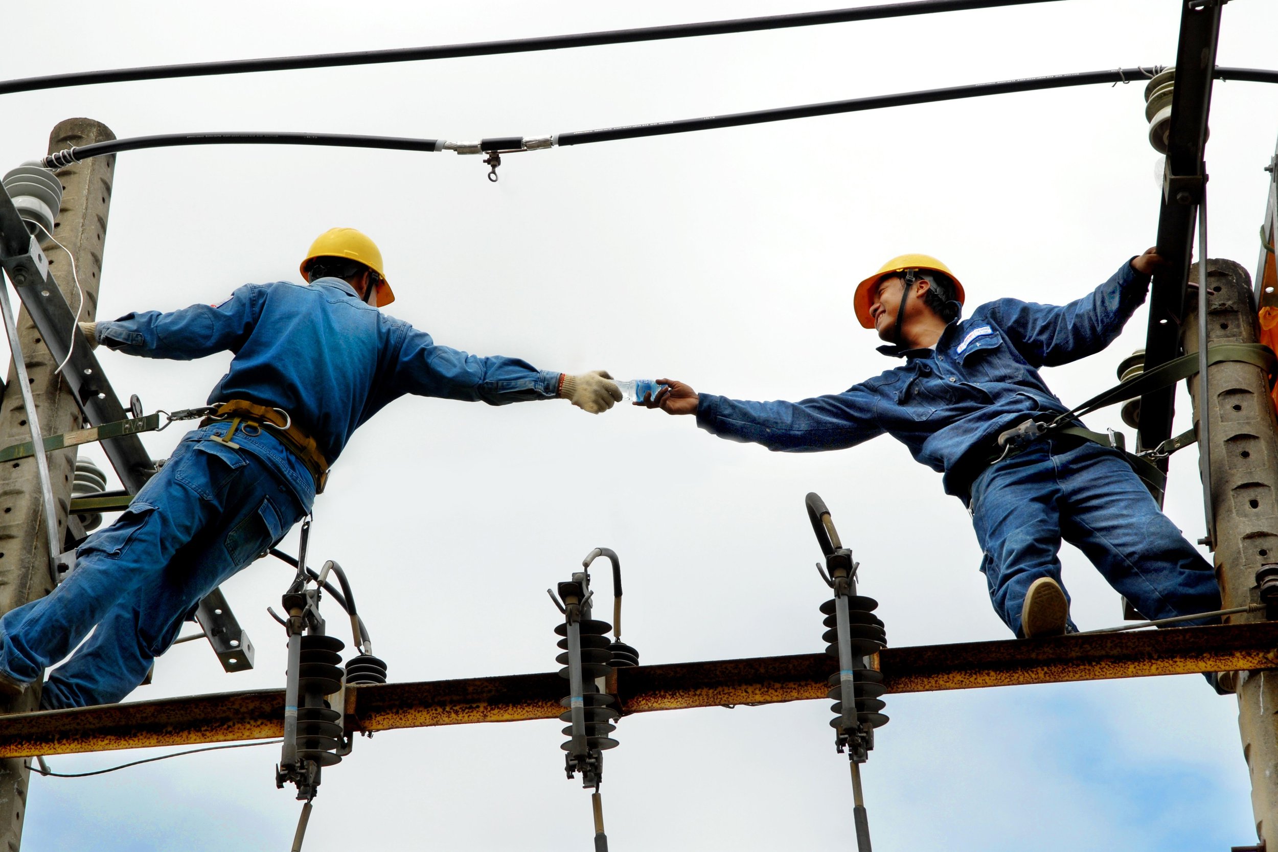 Dos trabajadores con cascos de seguridad trabajando en una estructura eléctrica, entregándose una botella de agua.
