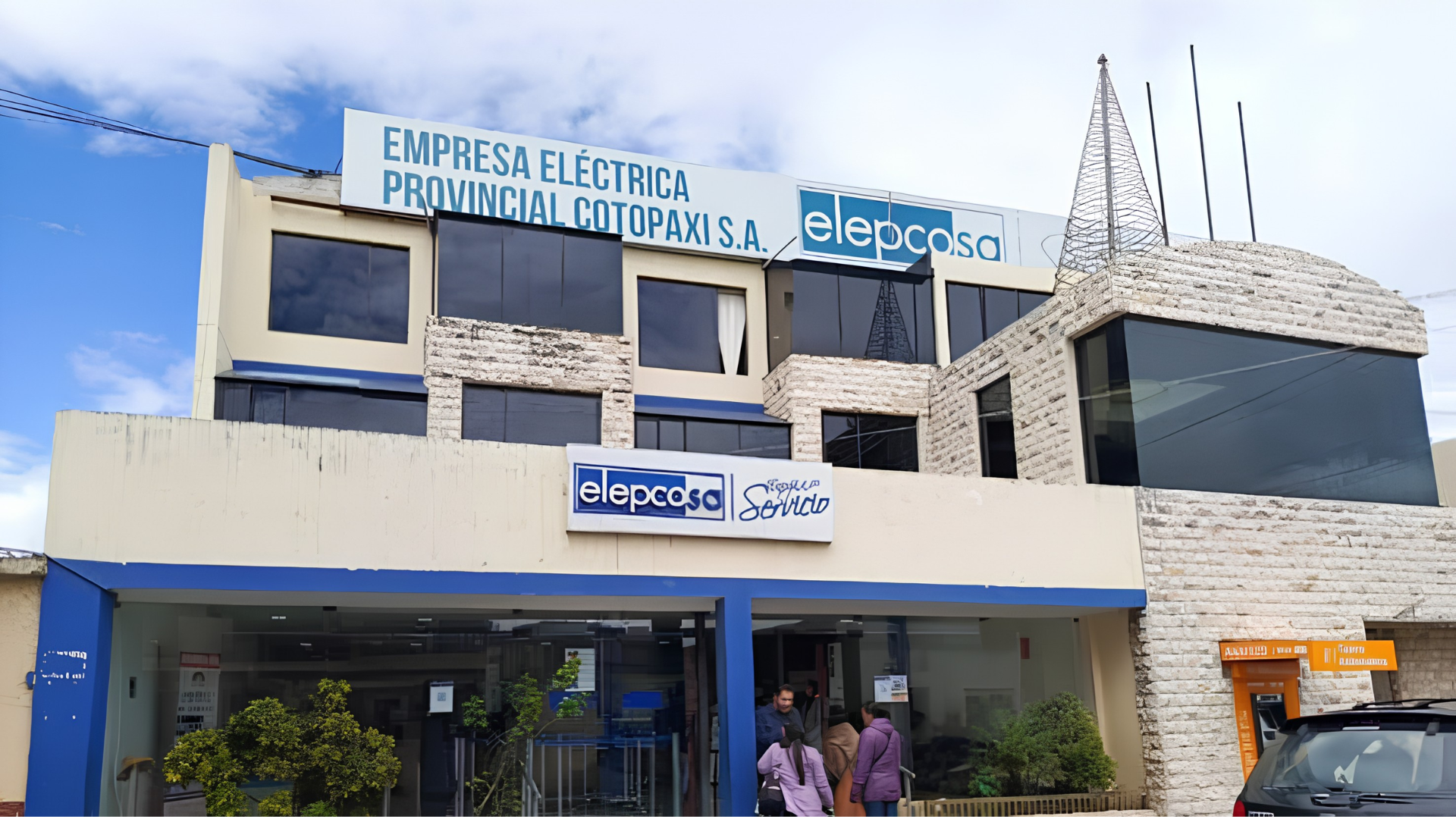 Edificio con señal de 'Empresa eléctrica provincial Cotopaxi S.A.' y logotipo de 'elecasa', fachada moderna con ventanas de vidrio y paredes de piedra, presencia de personas en la entrada y una estructura metálica en forma de espiral en la parte superior.