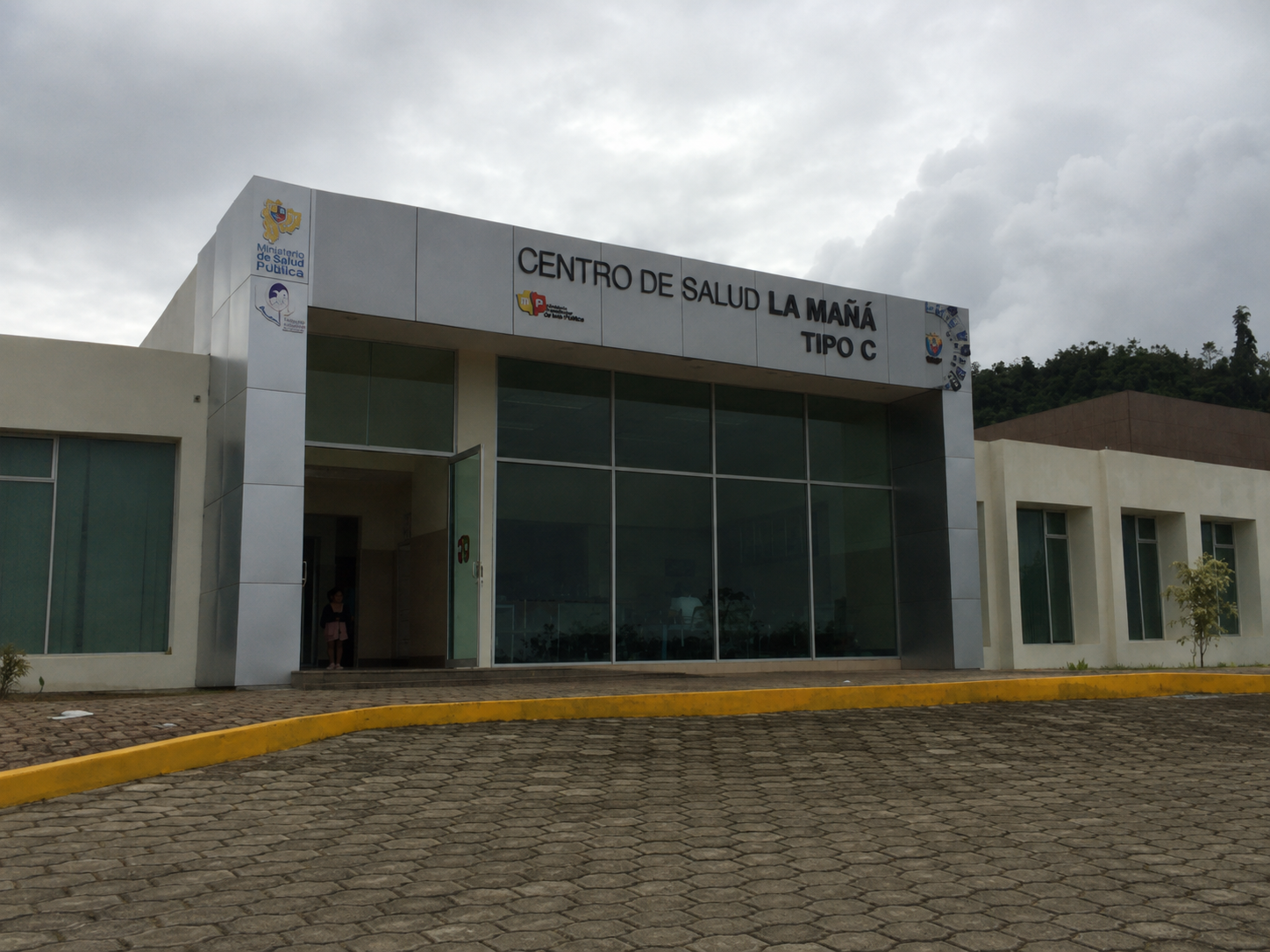 Fachada de un centro de salud llamado 'Centro de Salud La Mañá Tipo C' con logotipos del Ministerio de Salud Pública y del gobierno de México, sobre un pavimento de piedra y cielo nublado.