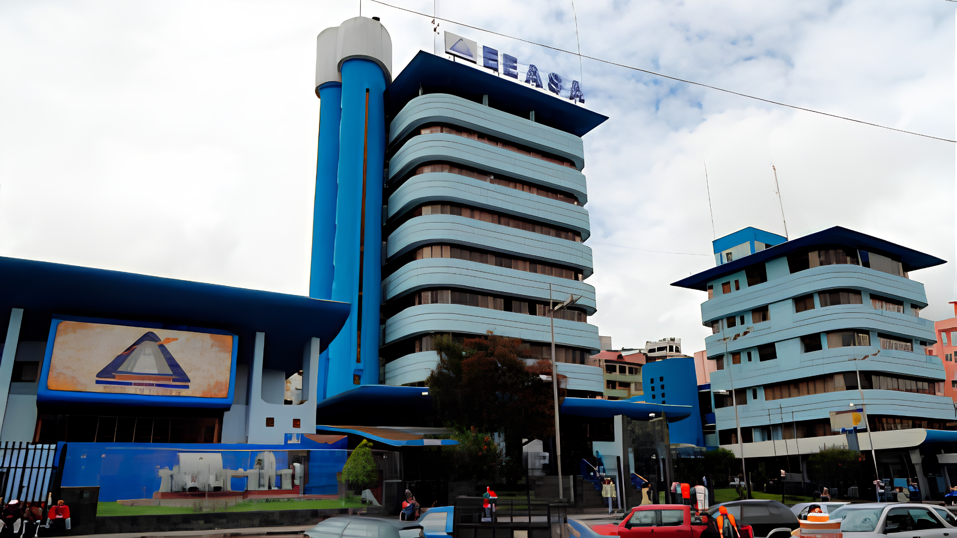 Edificio de arquitectura futurista en tonos azules y gris, con formas curvadas y varias plantas, en un ambiente urbano con vehículos y tren en primer plano, y cielo parcialmente nublado.