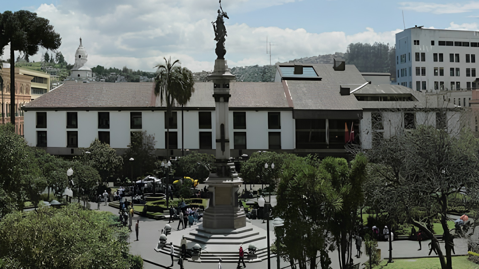 Una plaza céntrica con monumento en el centro, rodeada de árboles y edificios, muchas personas caminando y autos estacionados.