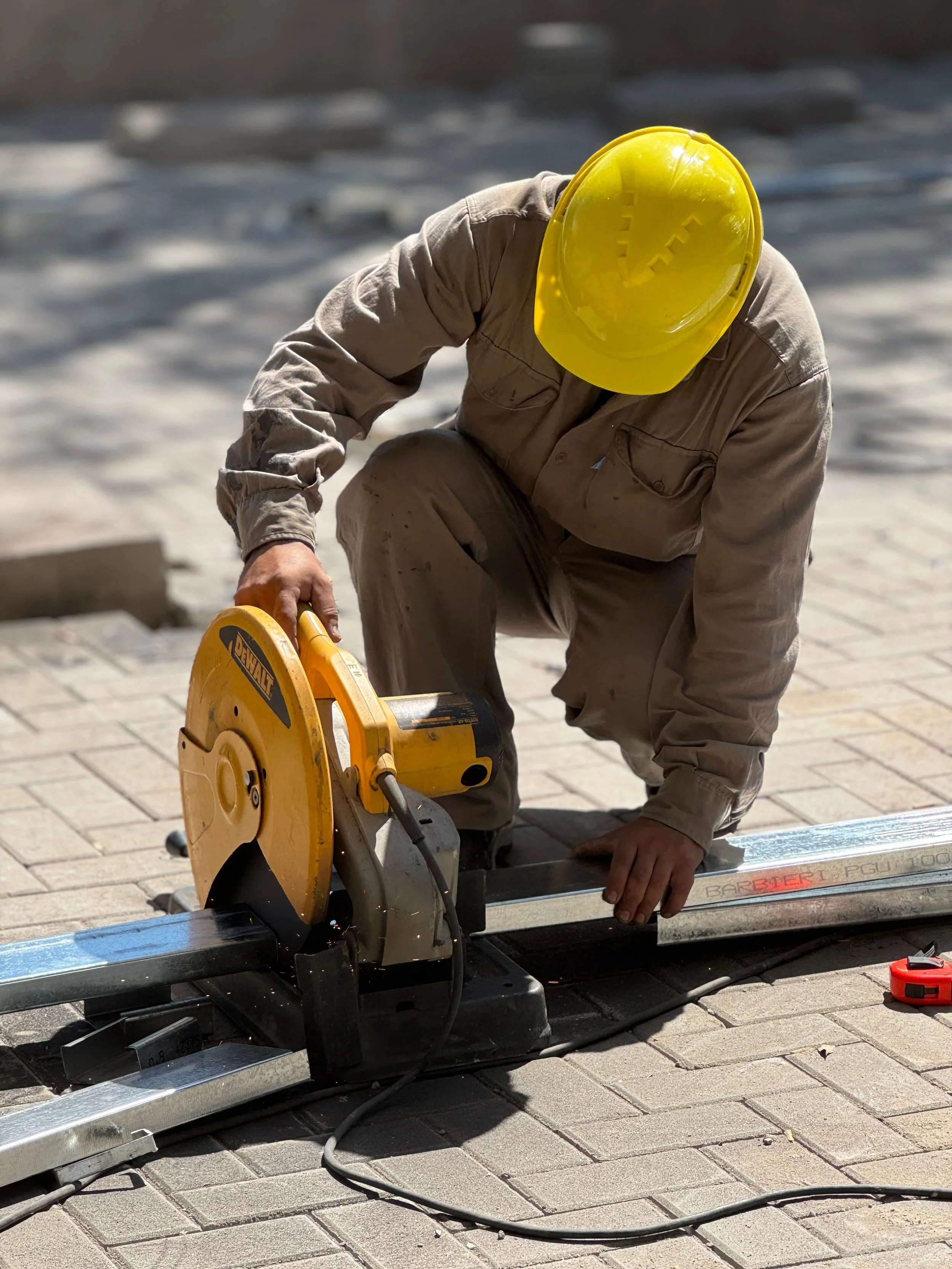 Un trabajador usando un casco amarillo y ropa de trabajo cortando una losa de piedra para pavimentar en un sitio de construcción.