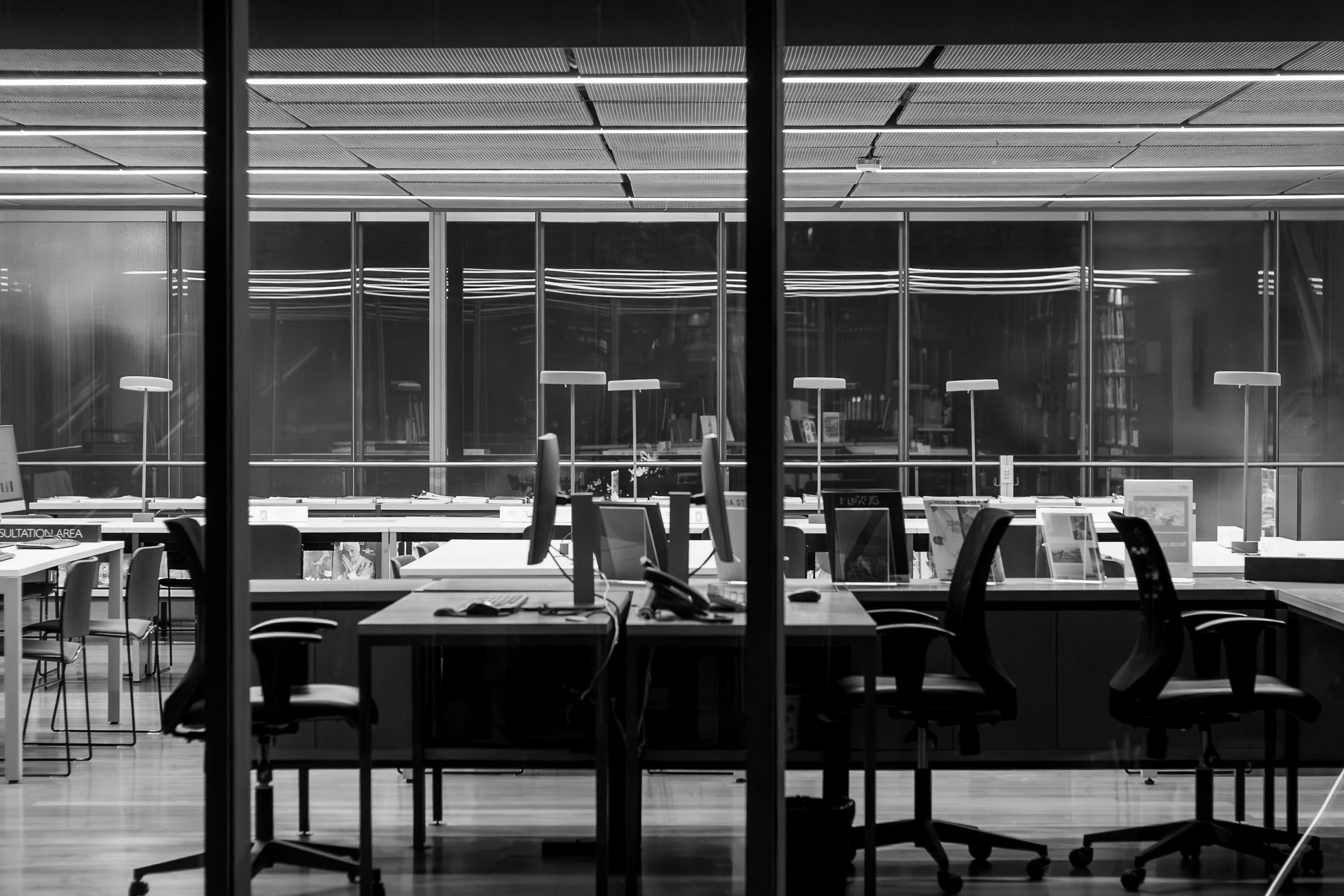 Oficina moderna con escritorios y computadoras, vista a través de ventanas de vidrio con cielo y edificios en la noche.