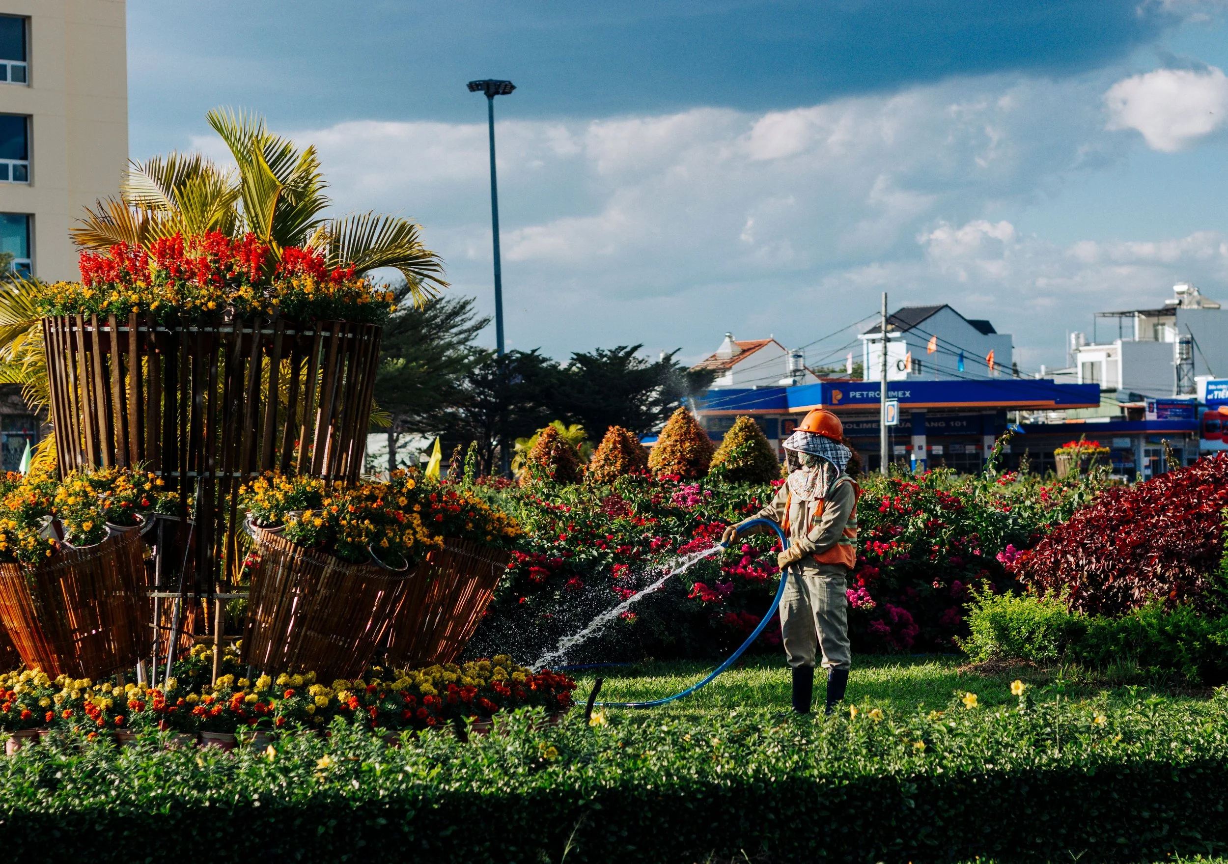Una persona cuidando un jardín de flores con una manguera. En el fondo se observa una gasolinera y edificios, cielo nublado.