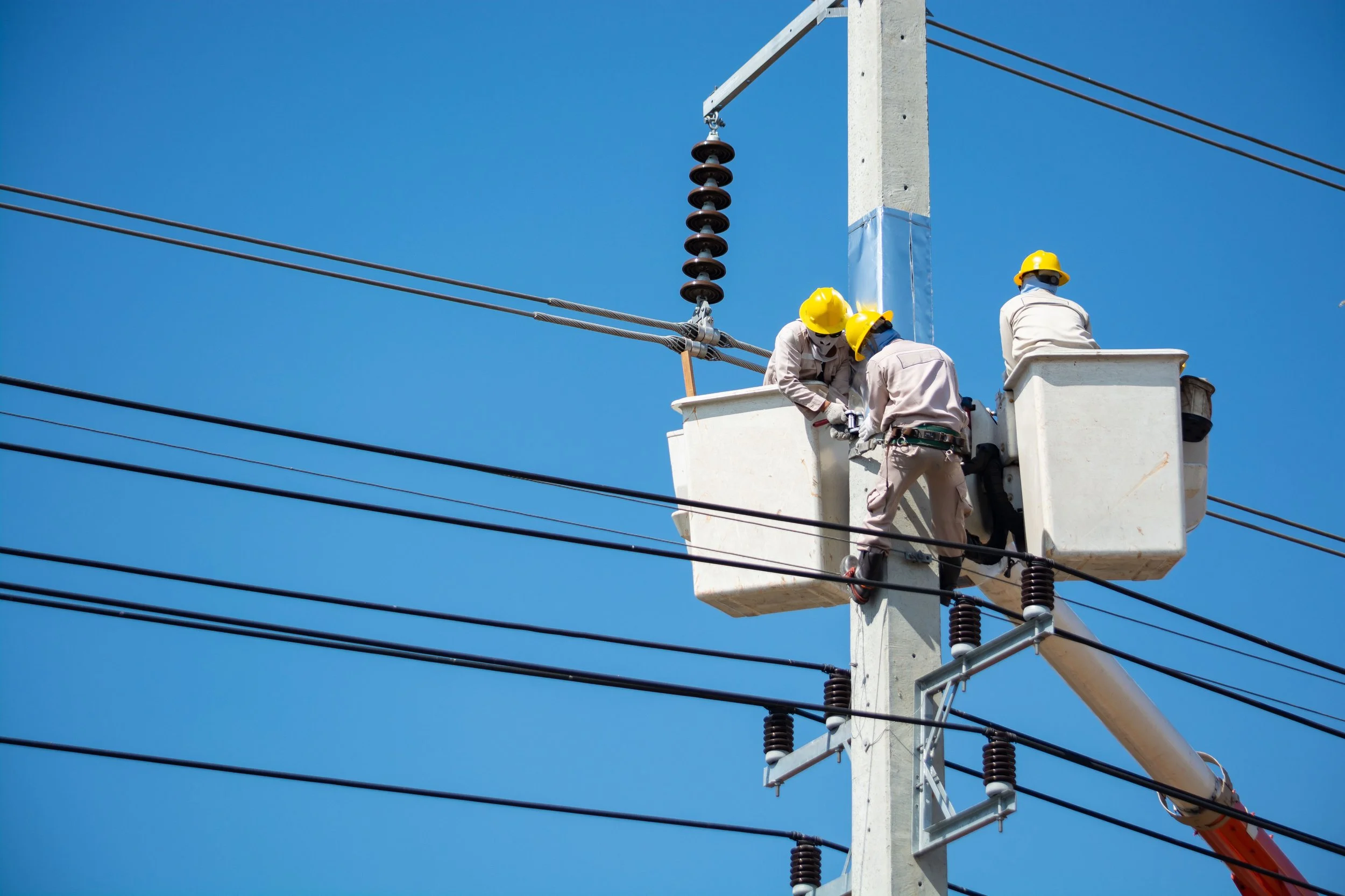 Trabajadores con cascos amarillos reparan líneas eléctricas en un poste, con cielo claro de fondo.