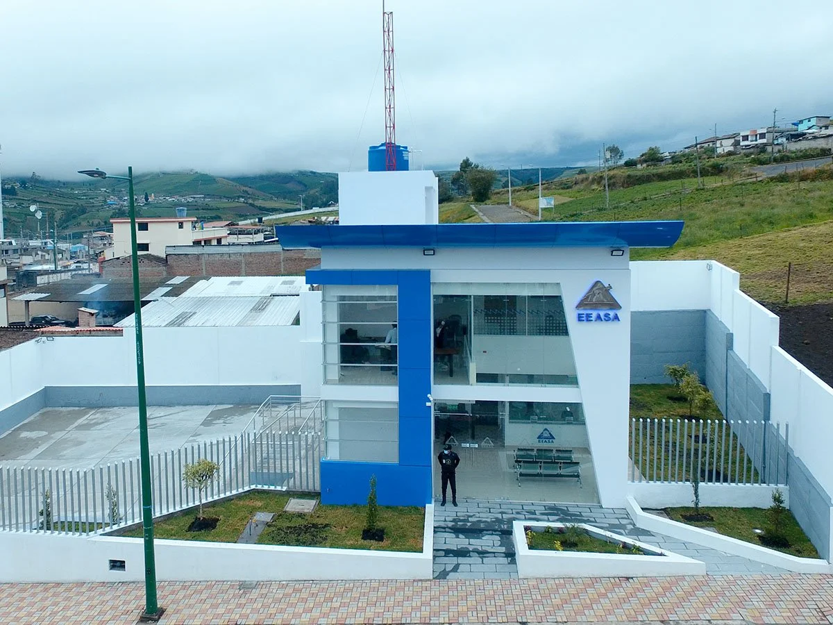 Edificio moderno con logotipo de EEASA, fachada de vidrio, en un entorno urbano con colinas y casas en el fondo.