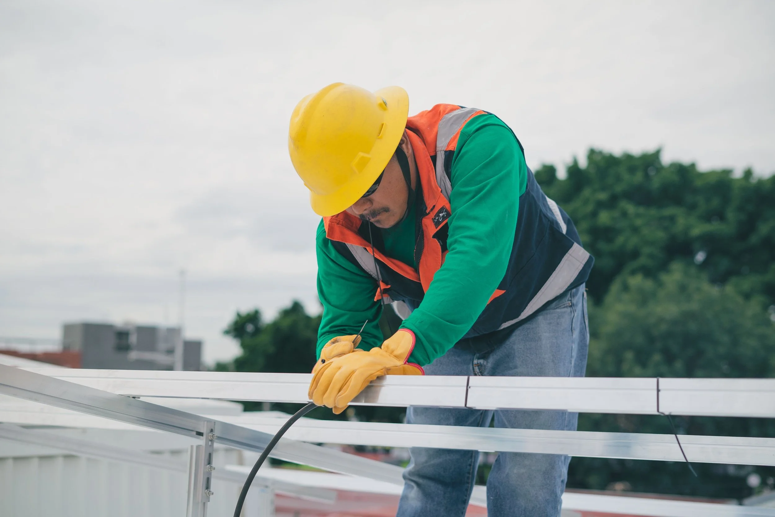 Un trabajador con casco amarillo, guantes amarillos, camiseta verde y chaleco de seguridad anaranjado trabaja en la instalación de paneles solares en un techo. El cielo está nublado y hay árboles y edificios en el fondo.