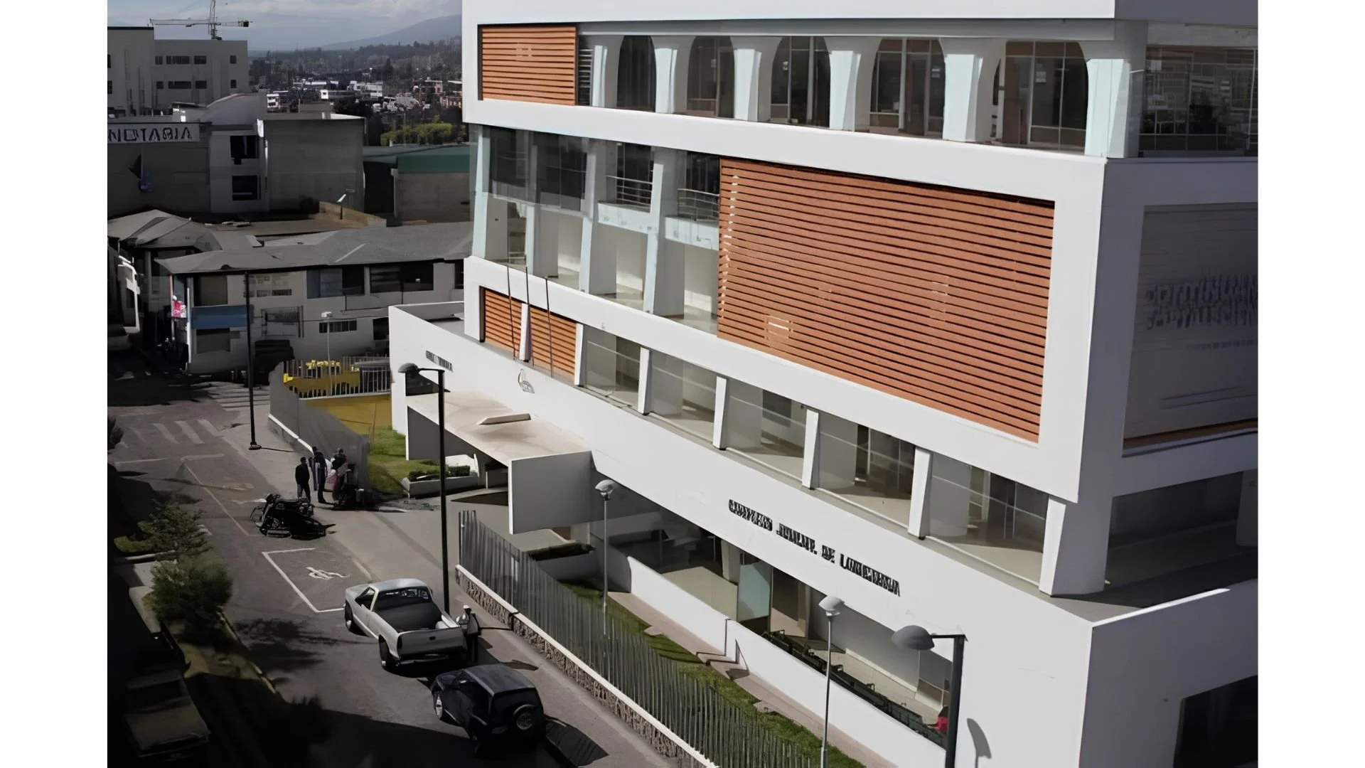 Edificio moderno de varios pisos con ventanas grandes y paneles de madera en las paredes externas, en un entorno urbano, con autos estacionados y personas caminando en la calle.