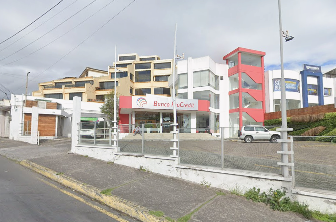 Edificio con fachada moderna que aloja un banco llamado Banco AroCredit, con varios autos y personas en el área de entrada y un muro con rejas metálicas delante.