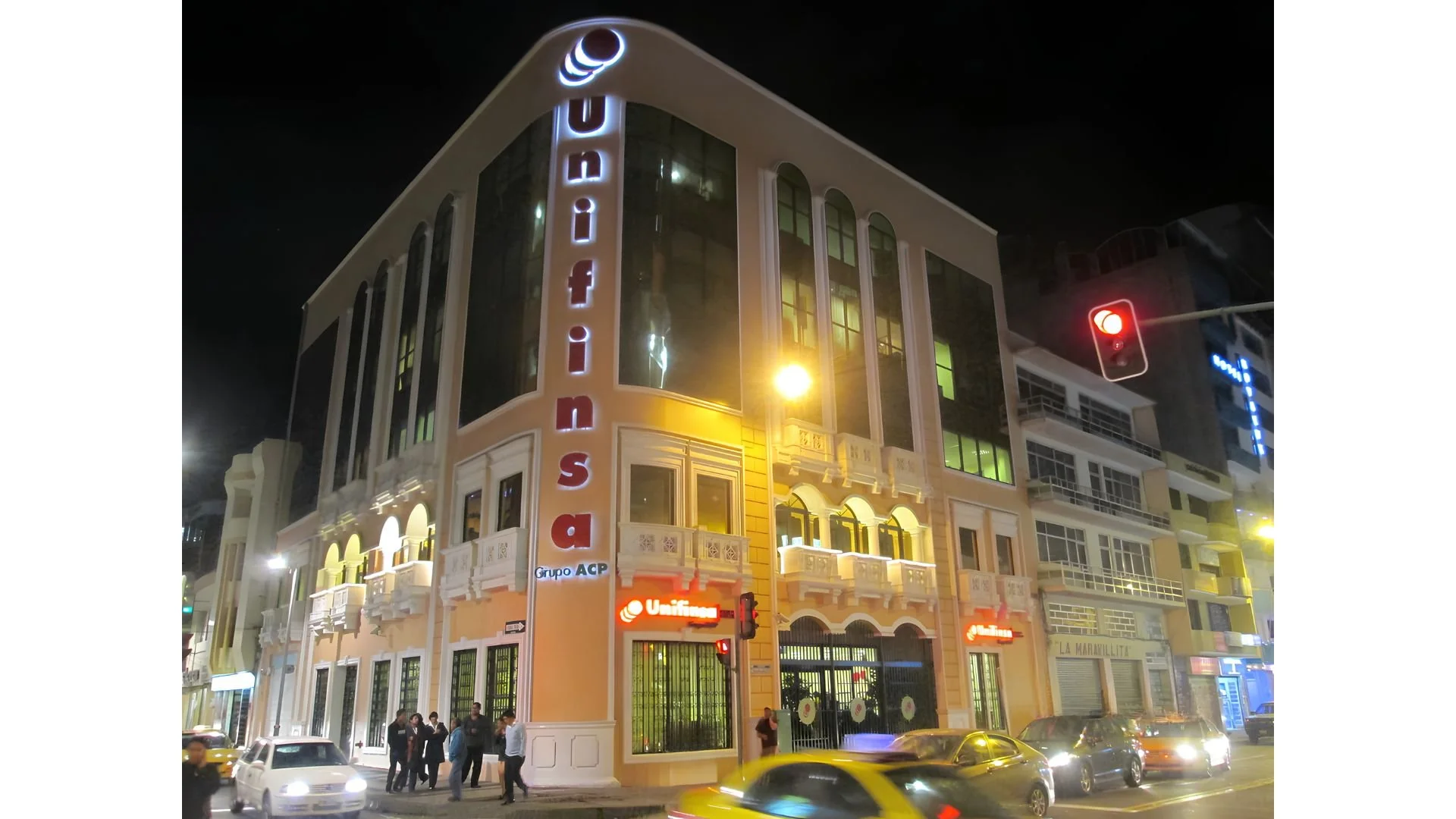 Edificio de varios pisos en la noche con letreros de neón que dicen 'UNIFIN' y 'grupo ACP', con personas y autos en la calle.