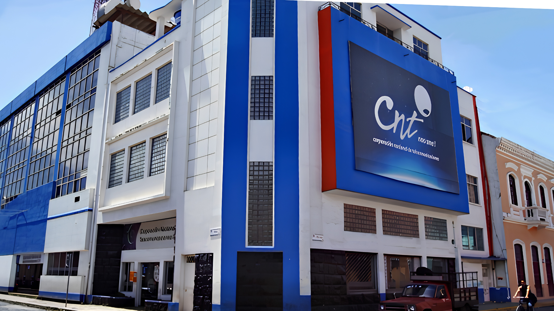 Edificio moderno de varios pisos con una pantalla digital que muestra el logo de CNT y sus redes sociales en la ciudad. Fachada en colores blanco, azul y rojo.