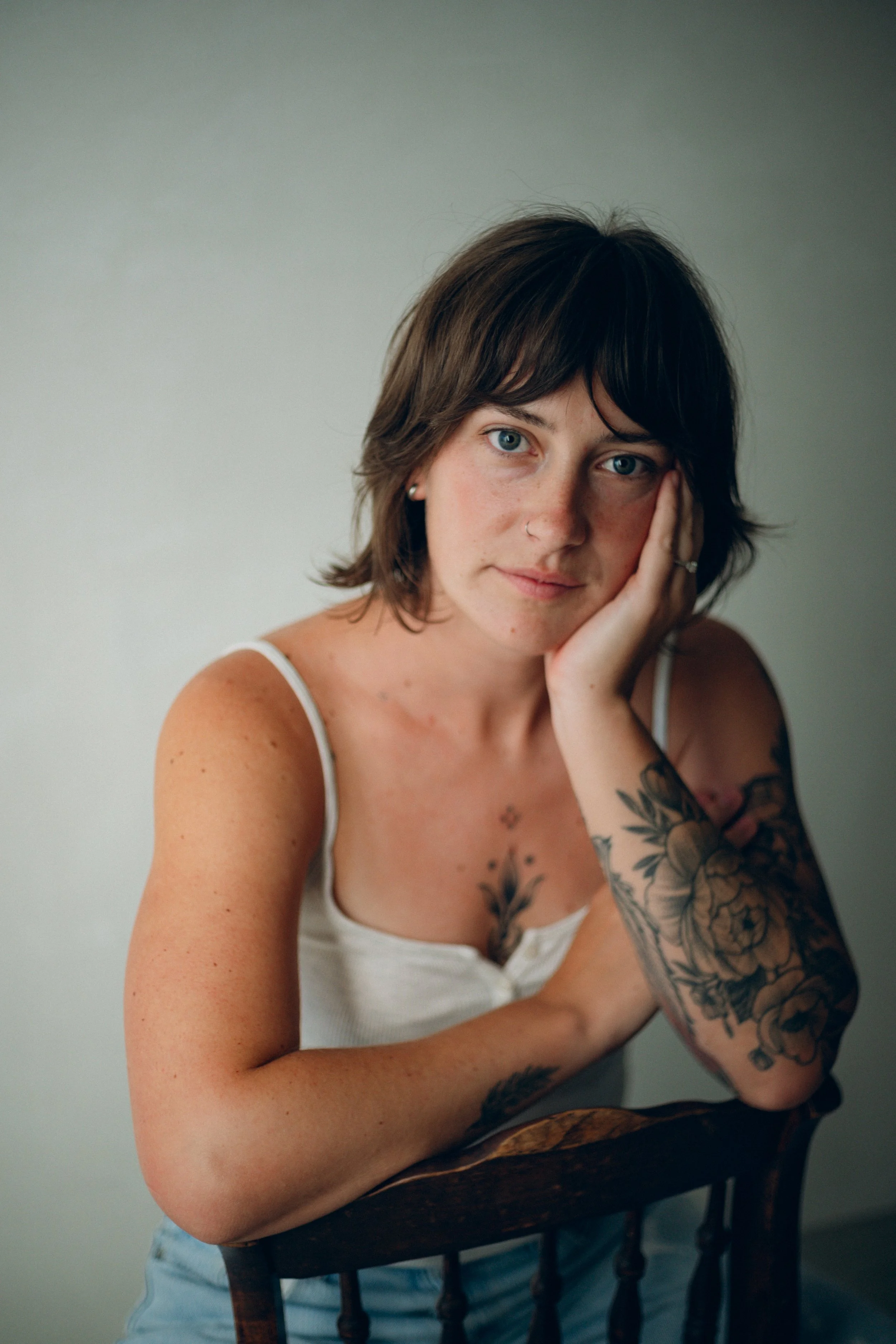 A woman with short brown hair, blue eyes, and tattoos on her arm, wearing a white tank top, sitting on a wooden chair with a neutral wall in the background.