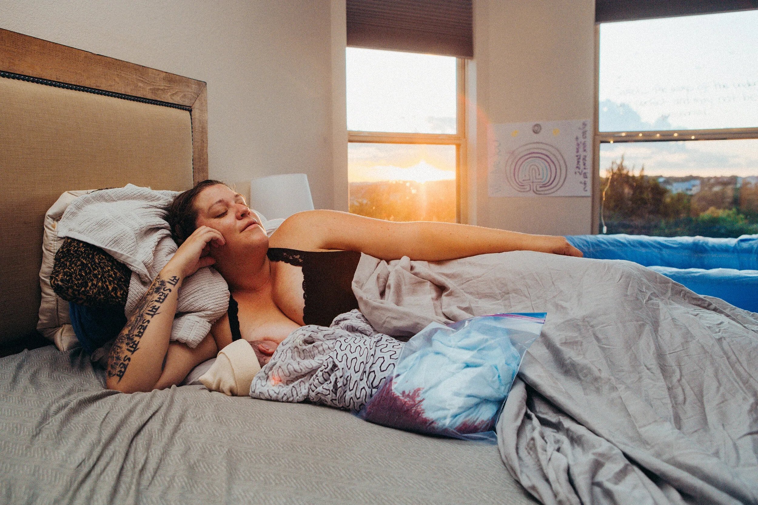 A woman with tattoos lying on a bed with her eyes closed, in a bedroom with sunlight streaming through windows.