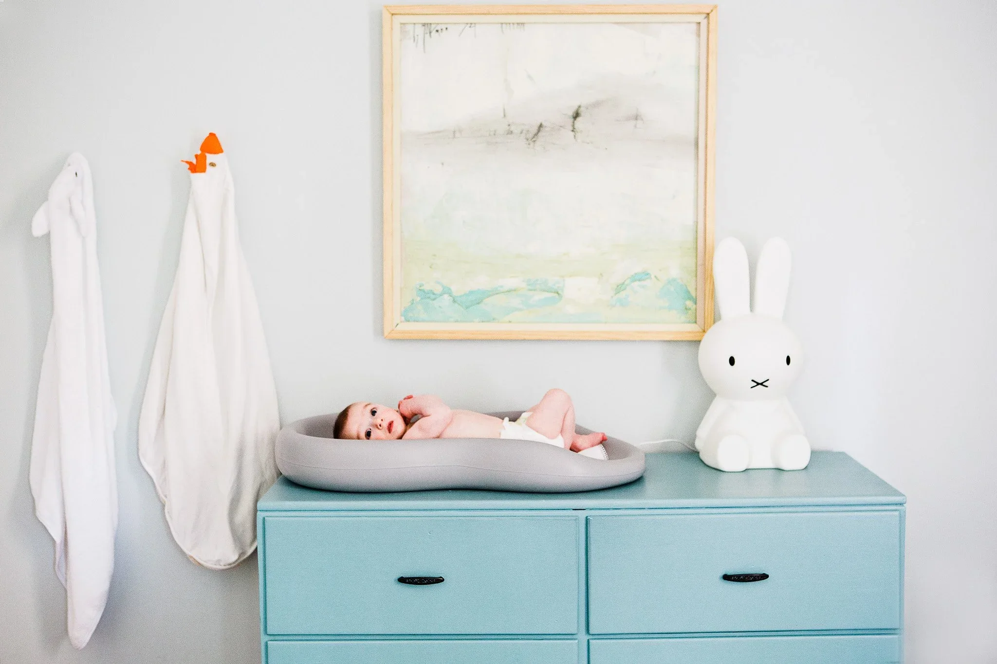 A baby lying in a gray nest on top of a light blue dresser, with white ghost and bunny plush toys on either side, and a framed light-colored landscape painting on the wall behind.